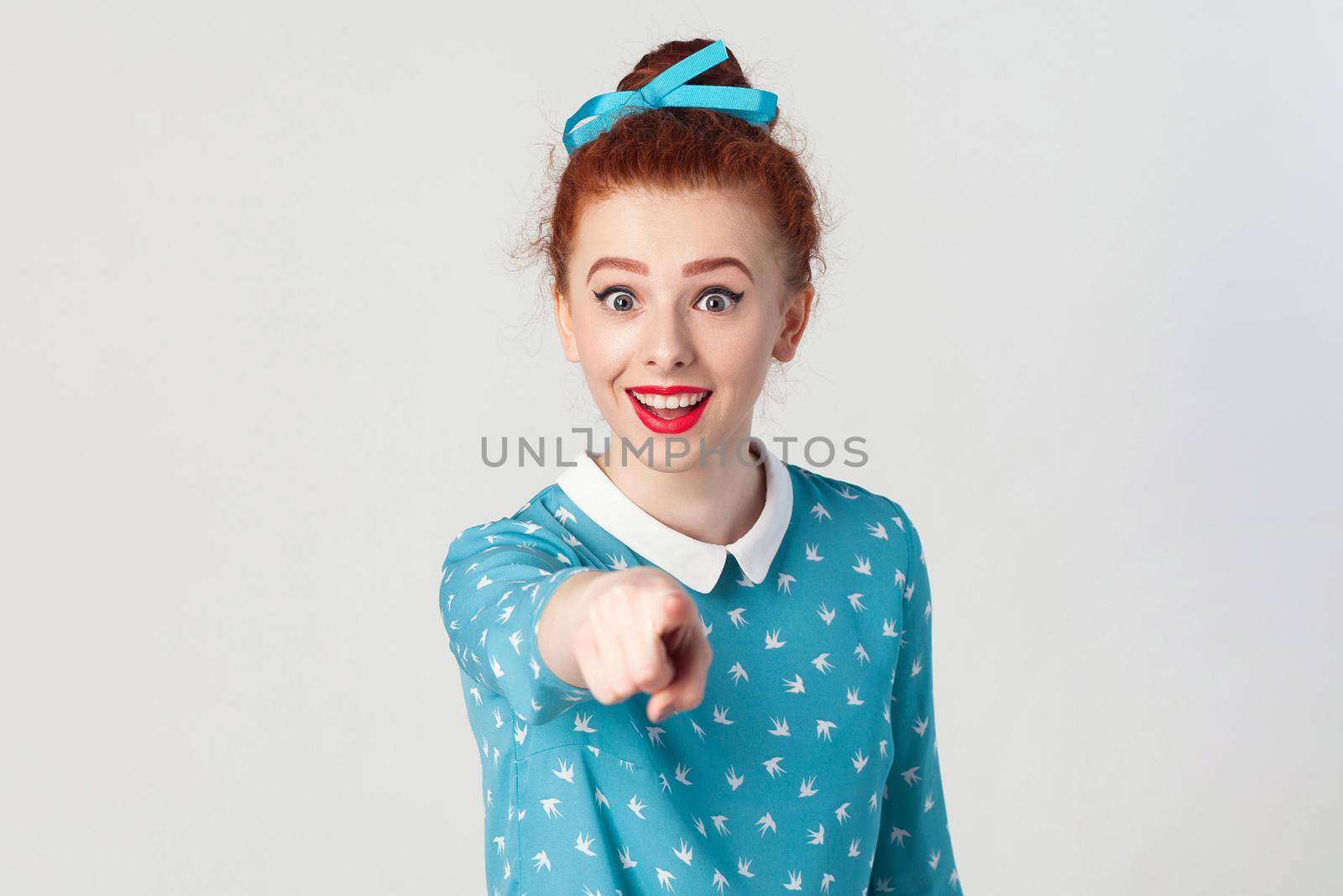 The beautiful redhead girl, wearing blue dress, opening mouths widely, having surprised shocked looks, pointing finger at camera. Isolated studio shot on gray background