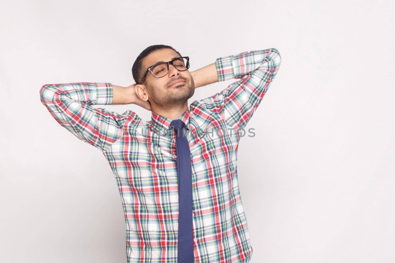 Portrait of dreamy handsome bearded businessman in checkered shirt, blue tie and eyeglasses standing with hand behind head and smiling with closed eyes. indoor studio shot, isolated on gray background