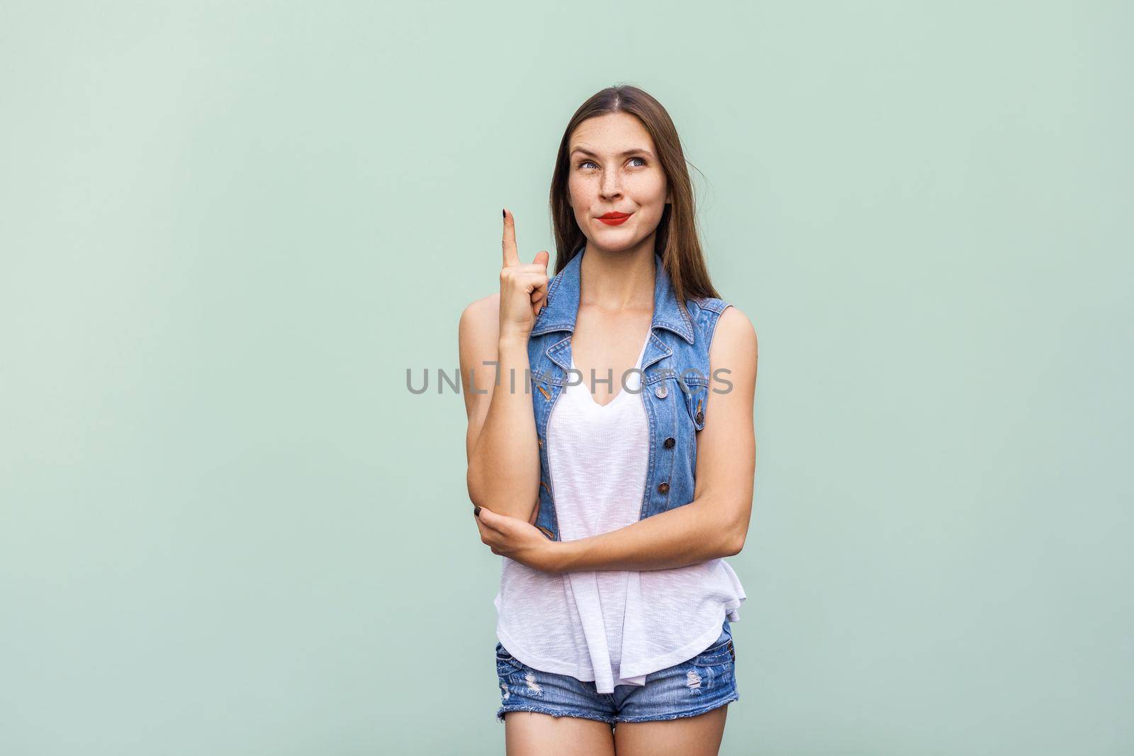 The tender and cute teenage female with shy faint smile, freckles and and clean skin, got the idea and she raised her finger up and looking up. Isolated studio shot on light green background