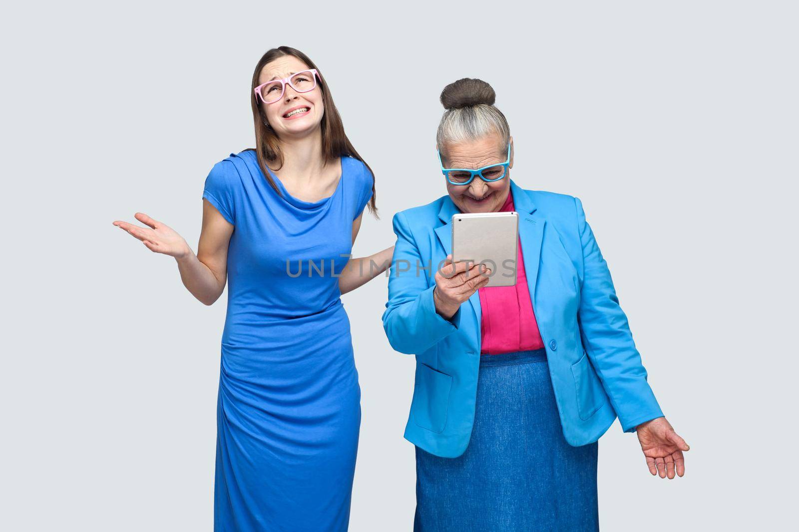 Why you did this? unhappy granddaughter woman standing near aged woman look in tablet. Relationship in the family. Granddaughter with grandmother. indoor, studio shot, isolated on gray background