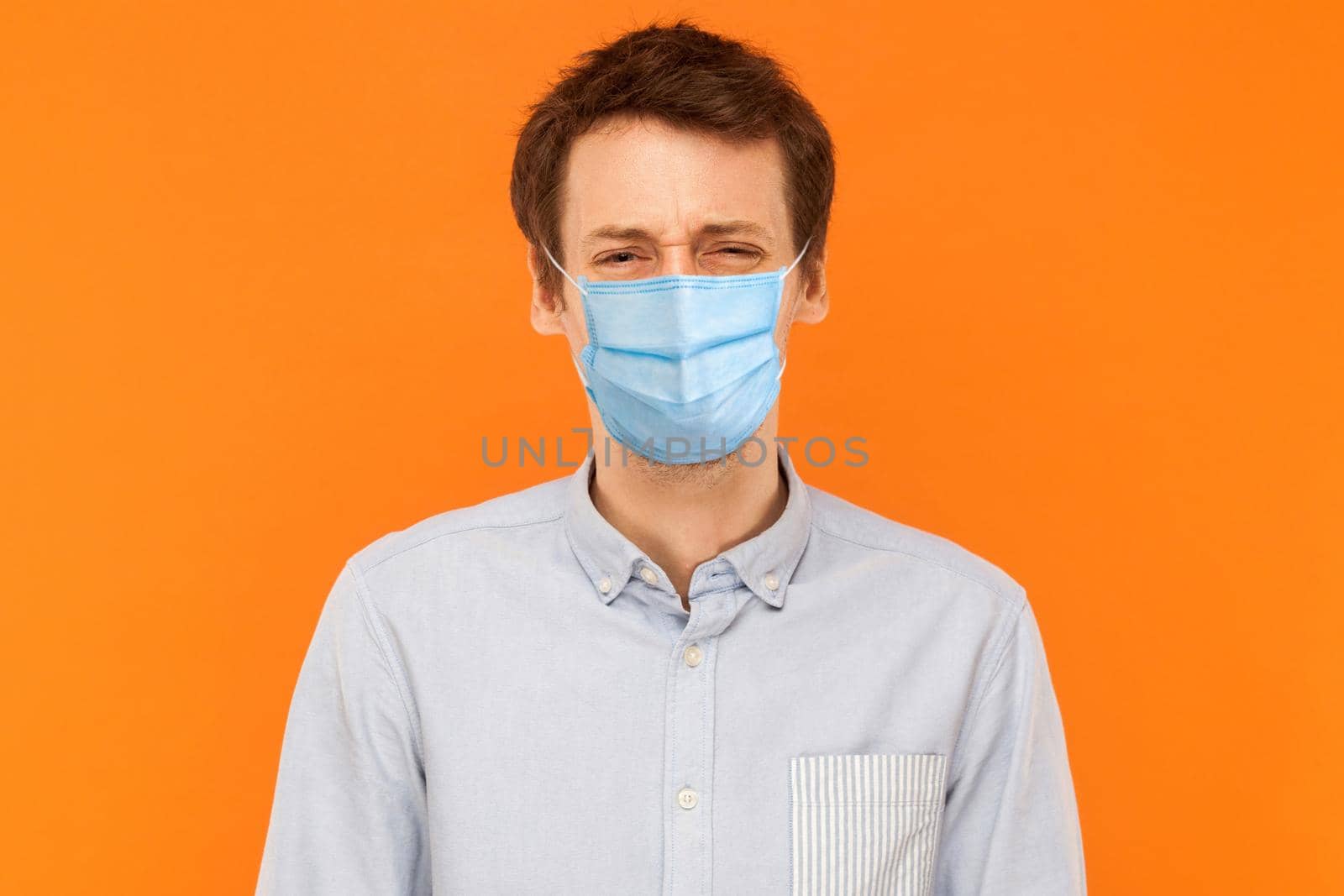 Portrait of sad alone young worker man with surgical medical mask standing and looking at camera with upset frown face. indoor studio shot isolated on orange background.