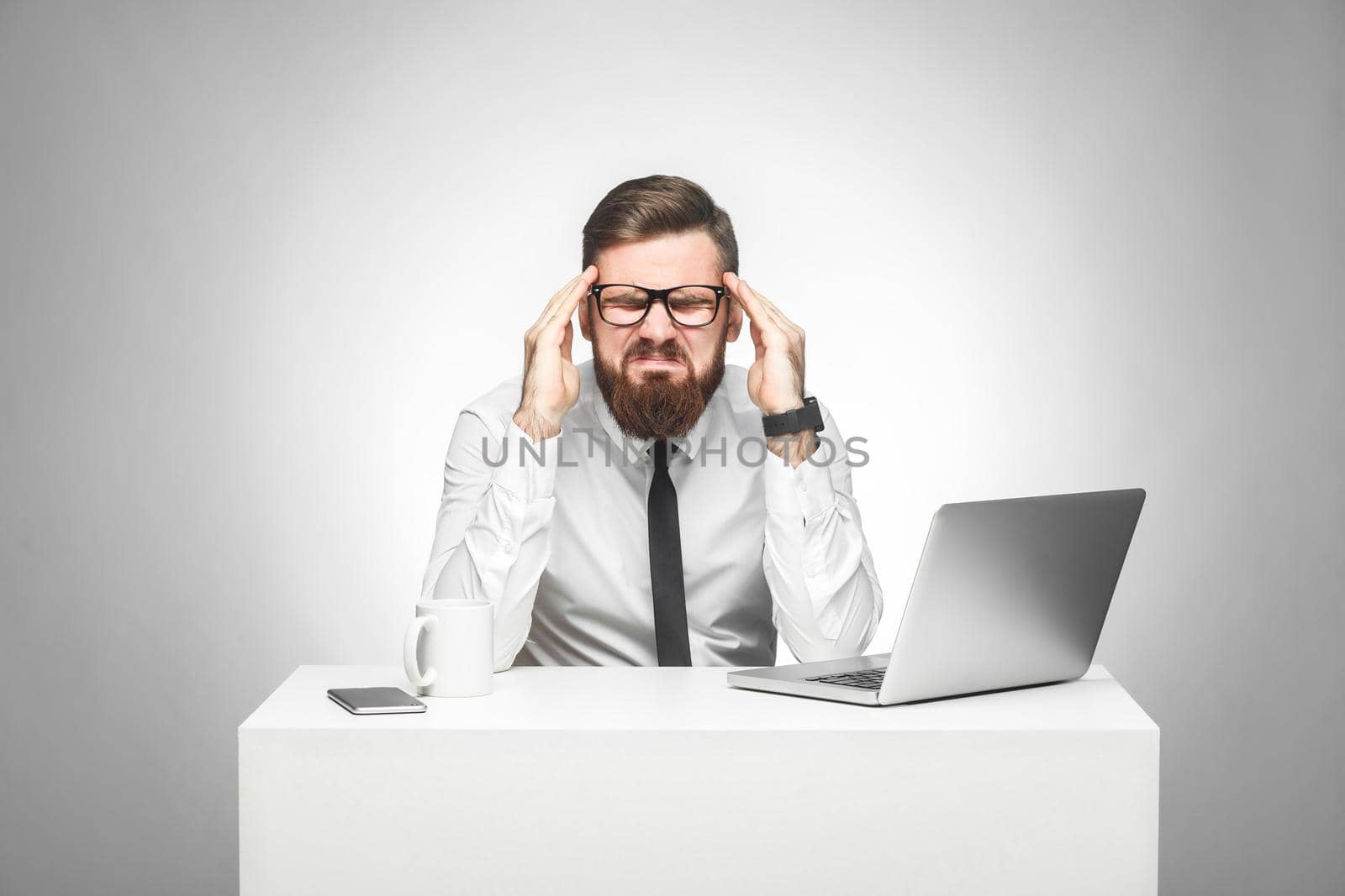 Portrait of emotional upset young manager in white shirt and black tie are sitting in office and grimacing cause made big mistake with stressed face, holding fingers near temples. Studio shot