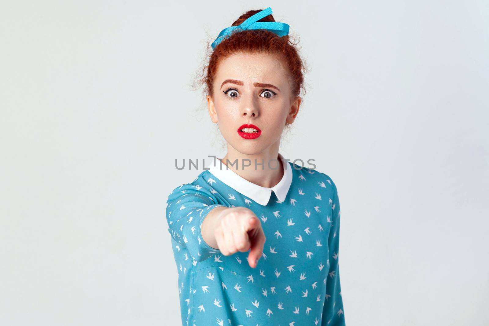 The beautiful caucasian girl, wearing blue dress, opening mouths widely, having surprised shocked looks, pointing finger at camera. Selective focus. Isolated studio shot on gray background