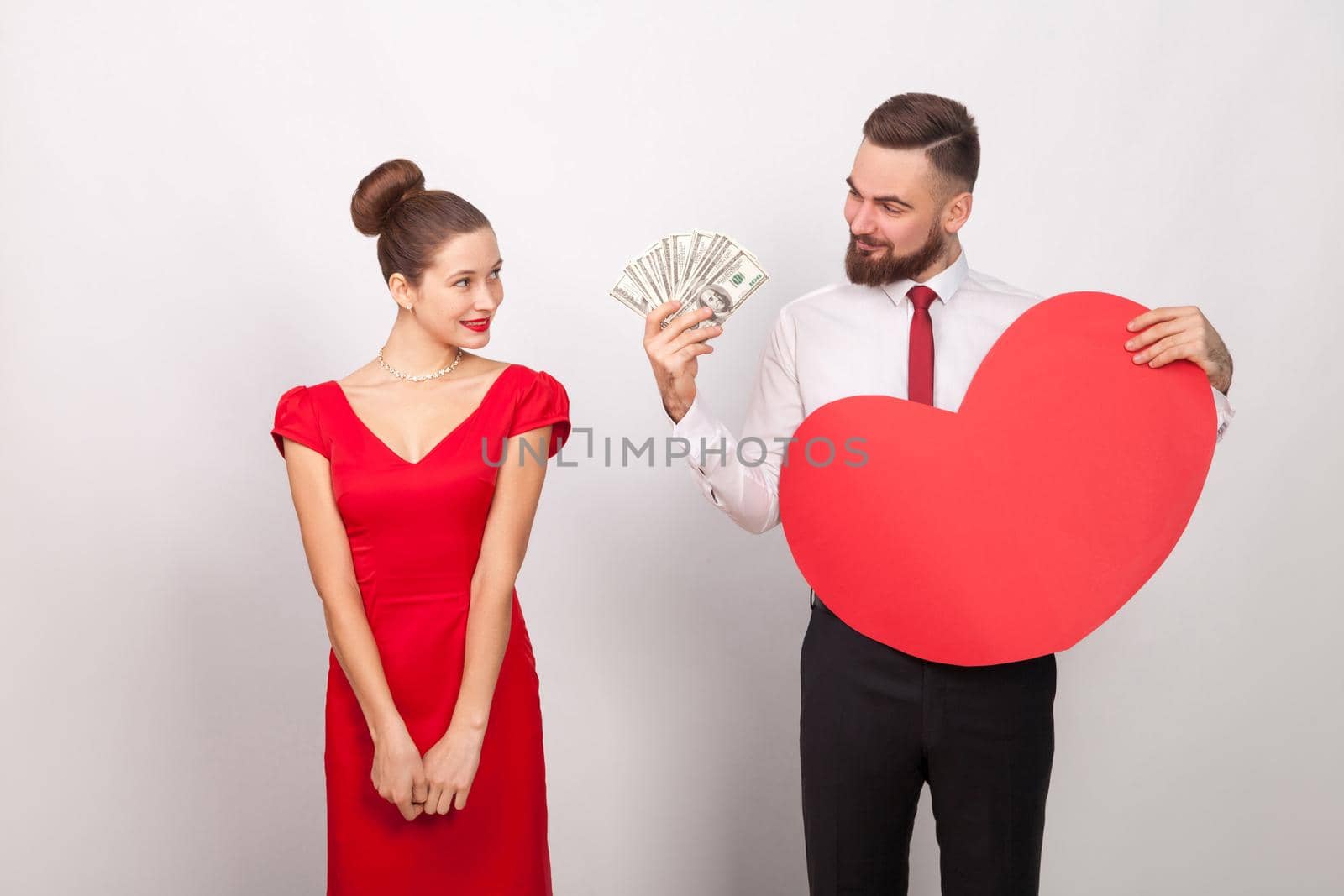 Business man holding many dollars and heart, cheerful woman embarrassed. Indoor, studio shot, isolated on gray background