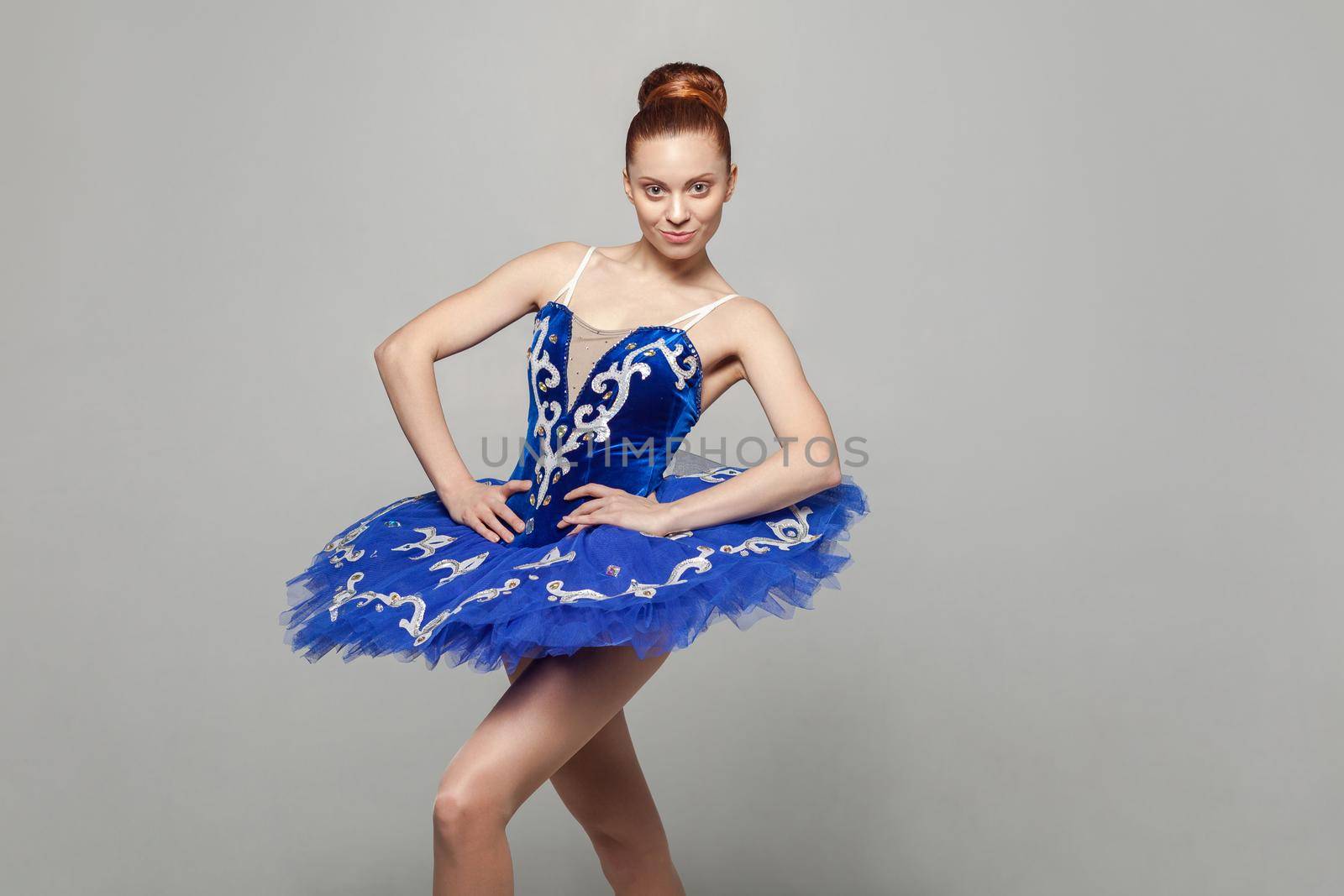 Portrait of beautiful happy ballerina woman in blue costume with makeup and bun collected hair standing against gray background, looking at camera. emotion and expression concept. indoor studio shot.