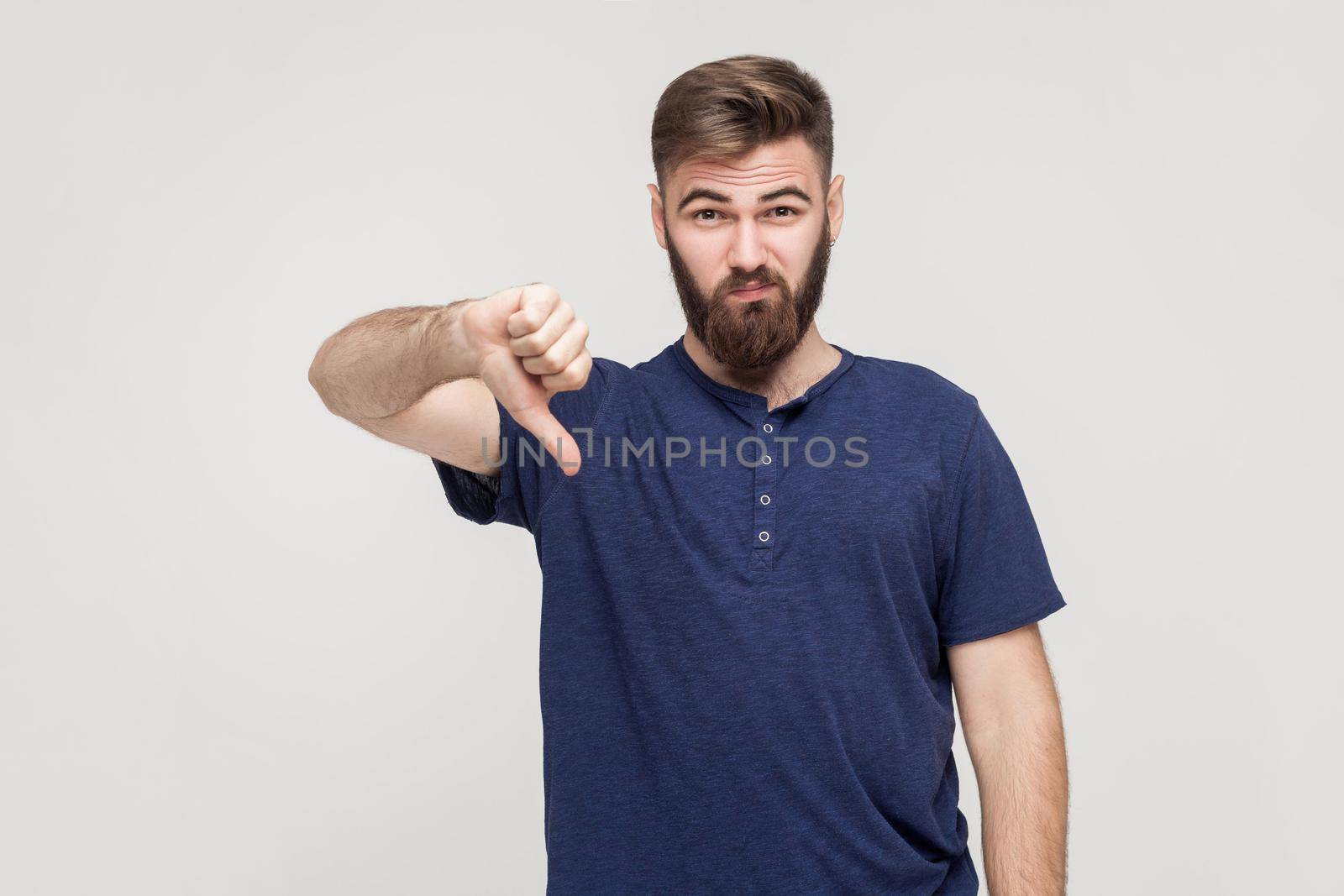 Portrait of unsatisfied bearded man with thumbs down and blue t-shirt against gray background. Studio shot.