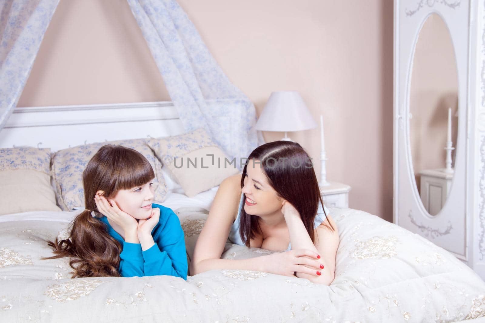 Happy mother and daughter lay in bed having fun, smiling and looking at camera. Studio shot.