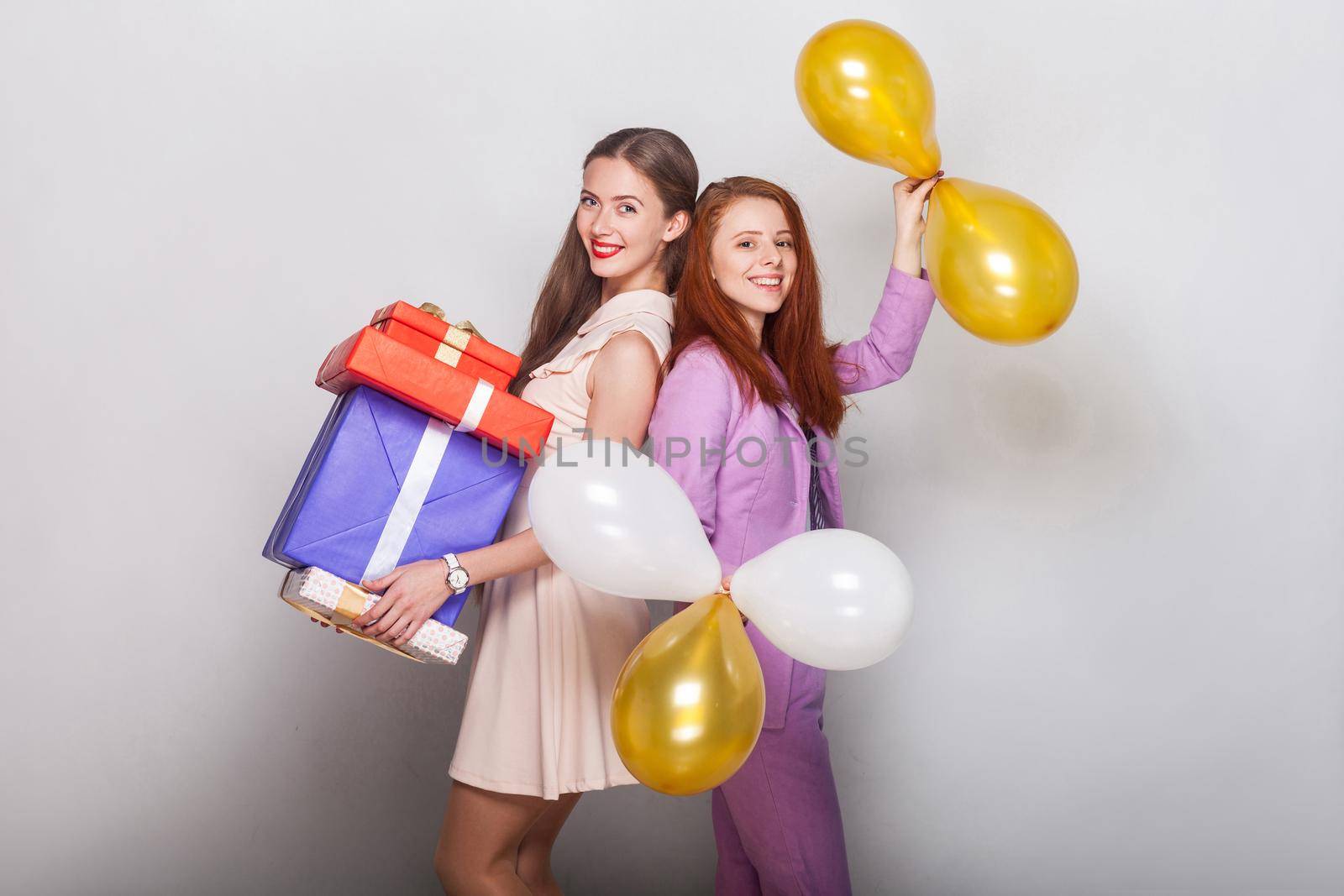 Beautiful girls standing back to back, holding many box and air balloon, toothy smiling and looking at camera. Studio shot, gray background