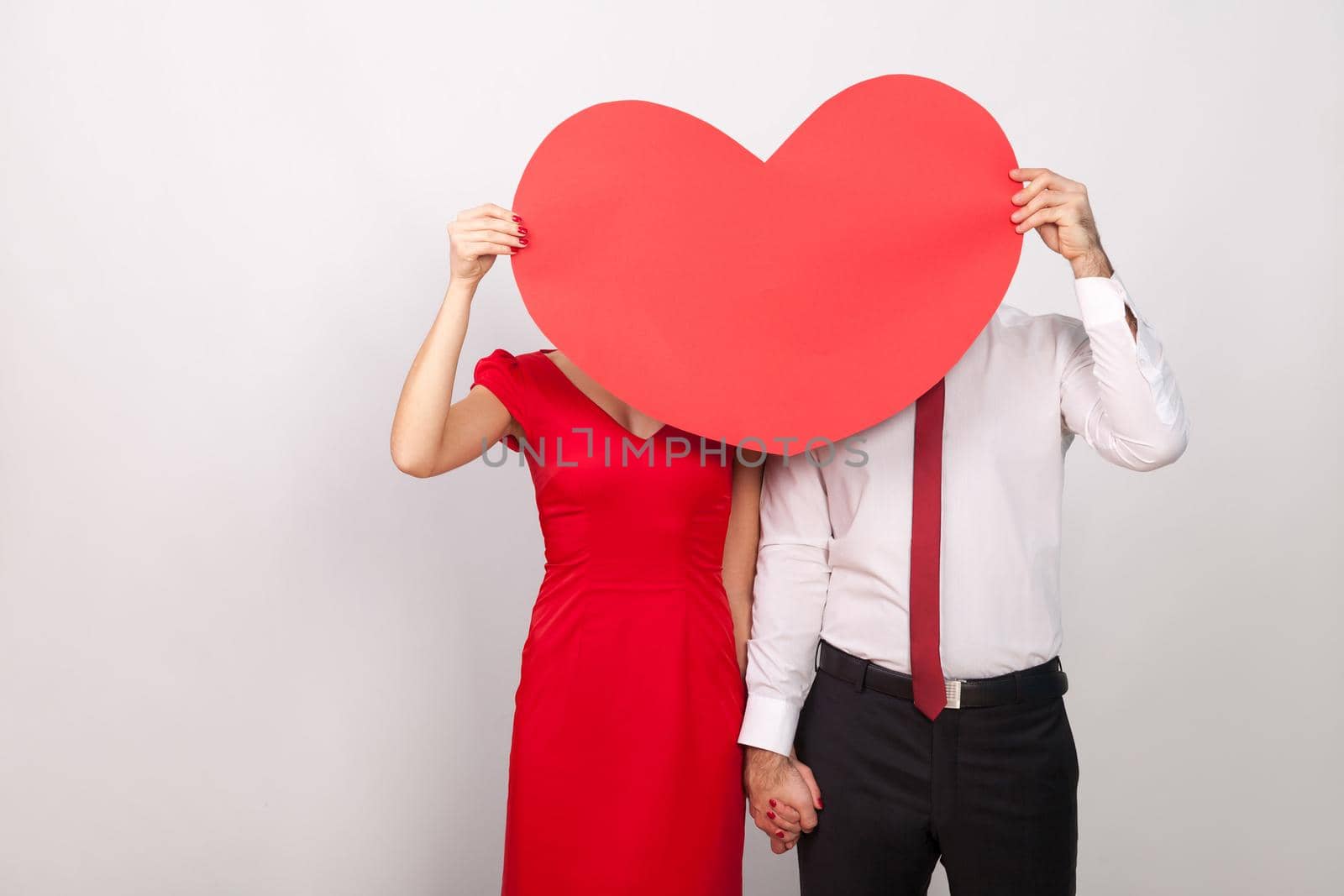 Unknown couple hide and seek behind big red heart. Indoor, studio shot, isolated on gray background