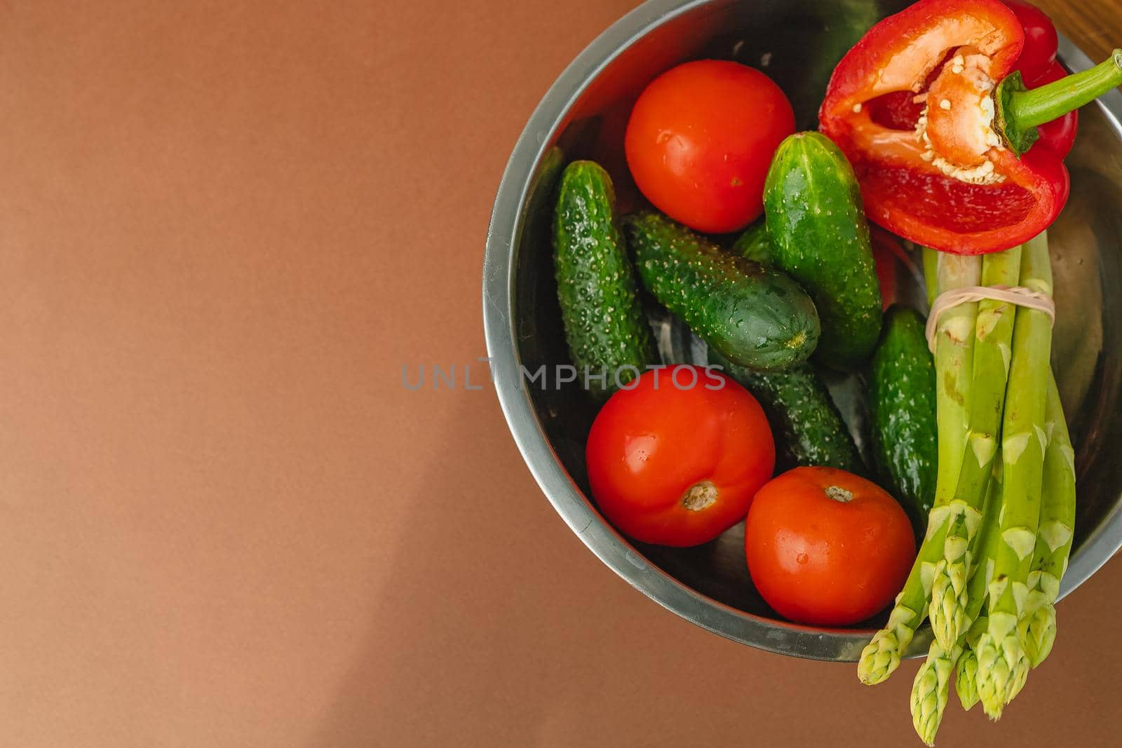 Vegetables lie in a metal bowl: tomatoes, asparagus, cucumbers, red bell peppers. on a wooden board and brown background. place for text. view from above.