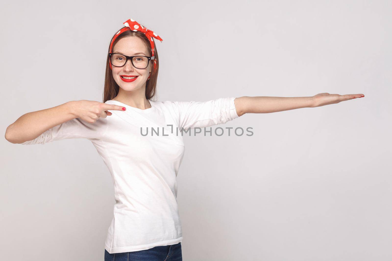 happy toothy smiley portrait of beautiful emotional young woman in white t-shirt with glasses, red lips and head band pointing and showing copy space. indoor studio shot, isolated on gray background.