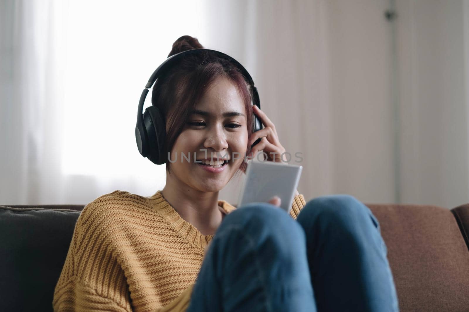 Attractive young woman on the sofa at home, she is playing music with her smarphone and wearing headphones, leisure and entertainment concept by wichayada