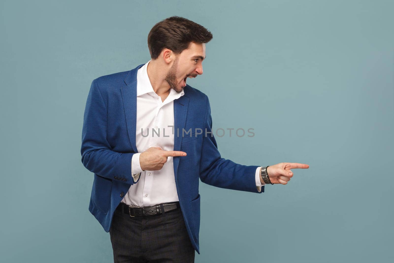Handsome amazed bearded man in blue suit standing and pointing at background and screaming and looking at copy space. studio, indoor shot isolated on blue background.