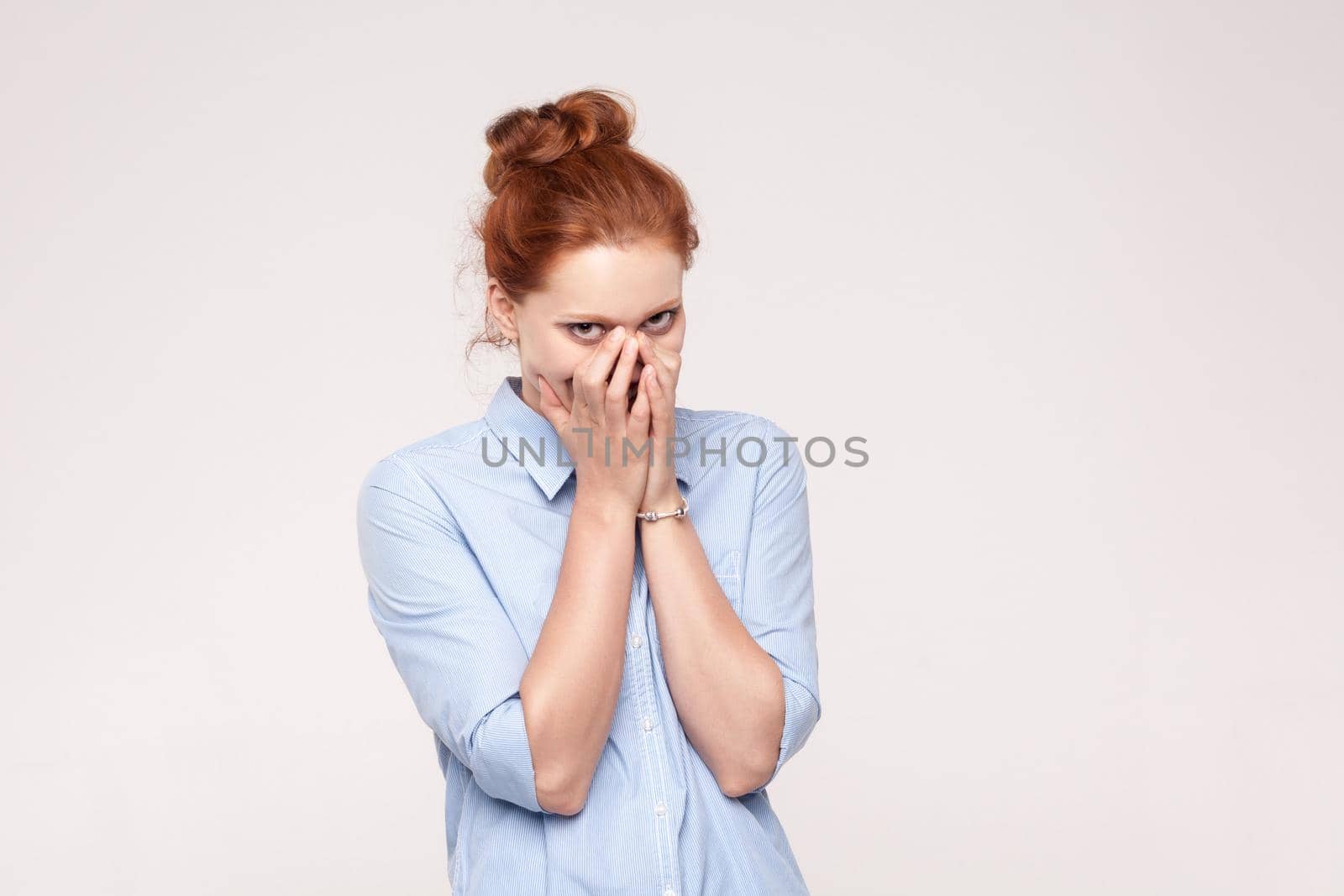 Shame and liar concept. Red haired woman covering mouth with both hands keeping a secret and little smiling. Beautiful redhead woman in blue shirt. Isolated studio shot on gray background.