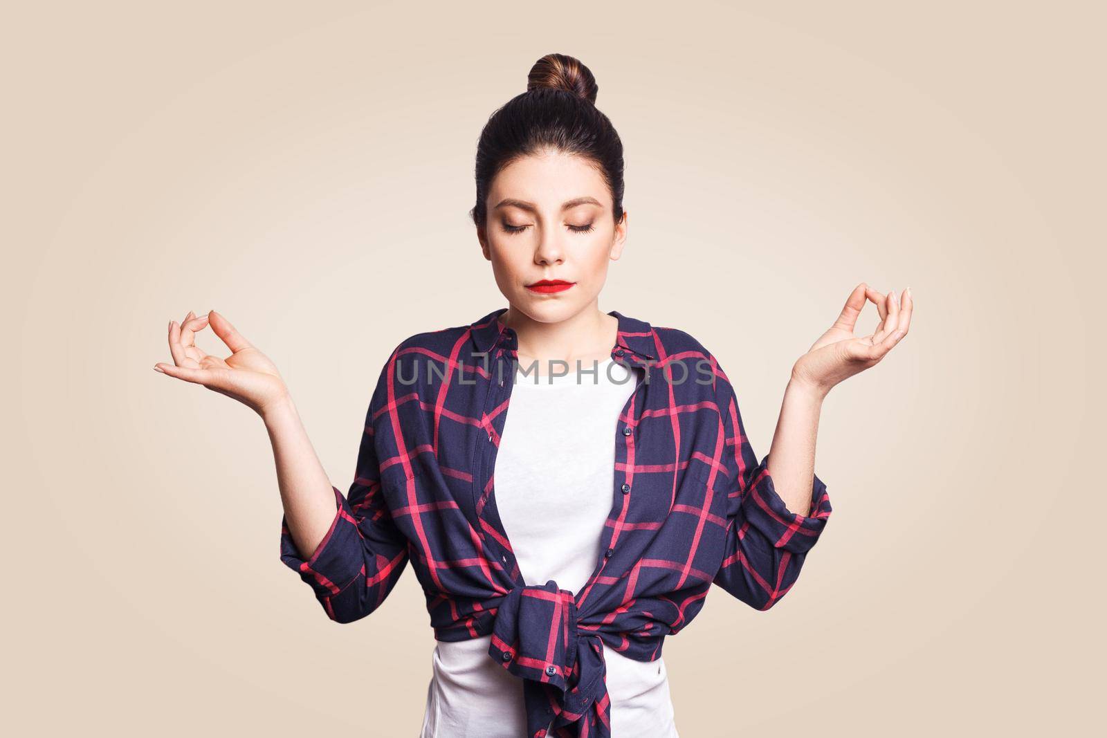 Meditation, religion and spiritual practises. Beautiful young brunette girl doing yoga in the morning indoors at beige wall after waking up, keeping eyes closed, holding fingers in mudra gesture