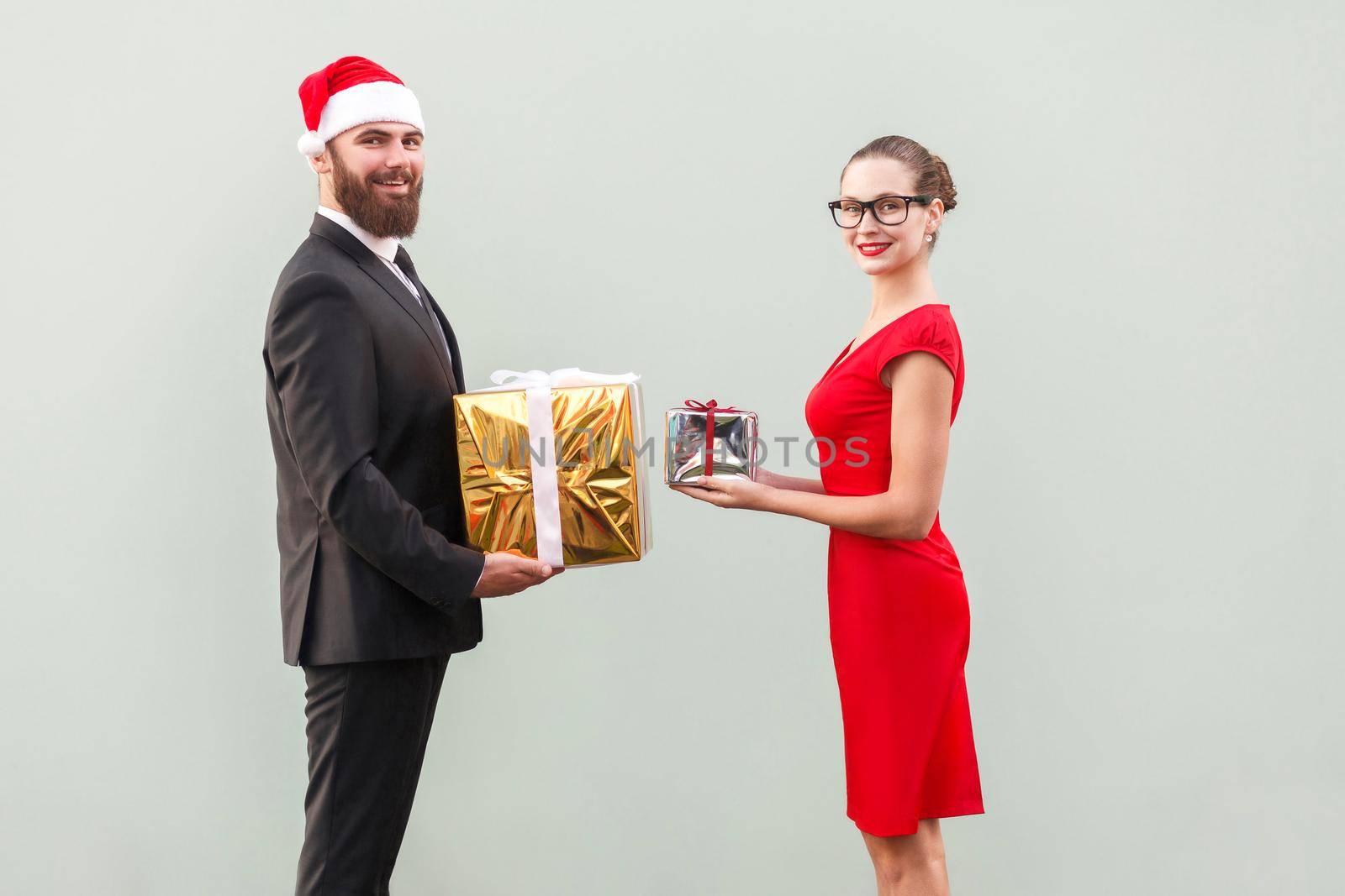 Happiness couple looking at camera with toothy smiling and give each other gift box, in honor of christmas. Studio shot. Isolated on gray background