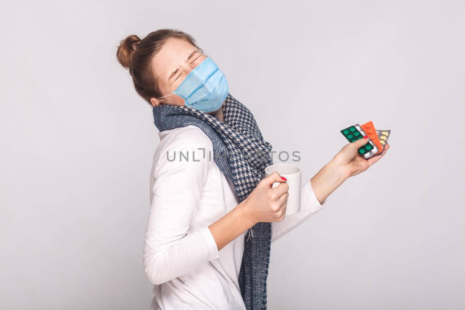 Woman with surgical medical mask, cry because she was sick. Holding cup with tea, many pills and antibiotics. indoor, studio shot, isolated on gray background