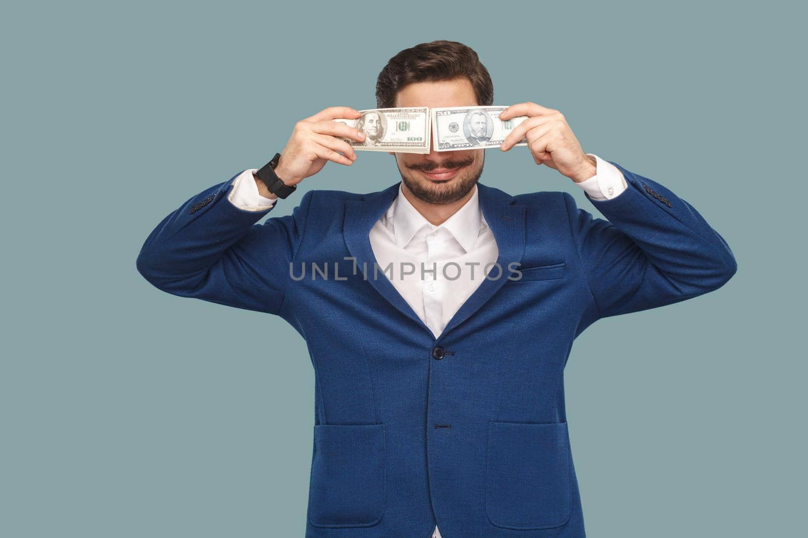 Handsome happy businessman in blue jacket and white shirt standing and holding and covered many dollars in front of eyes and smiling . Indoor, studio shot isolated on light blue background.