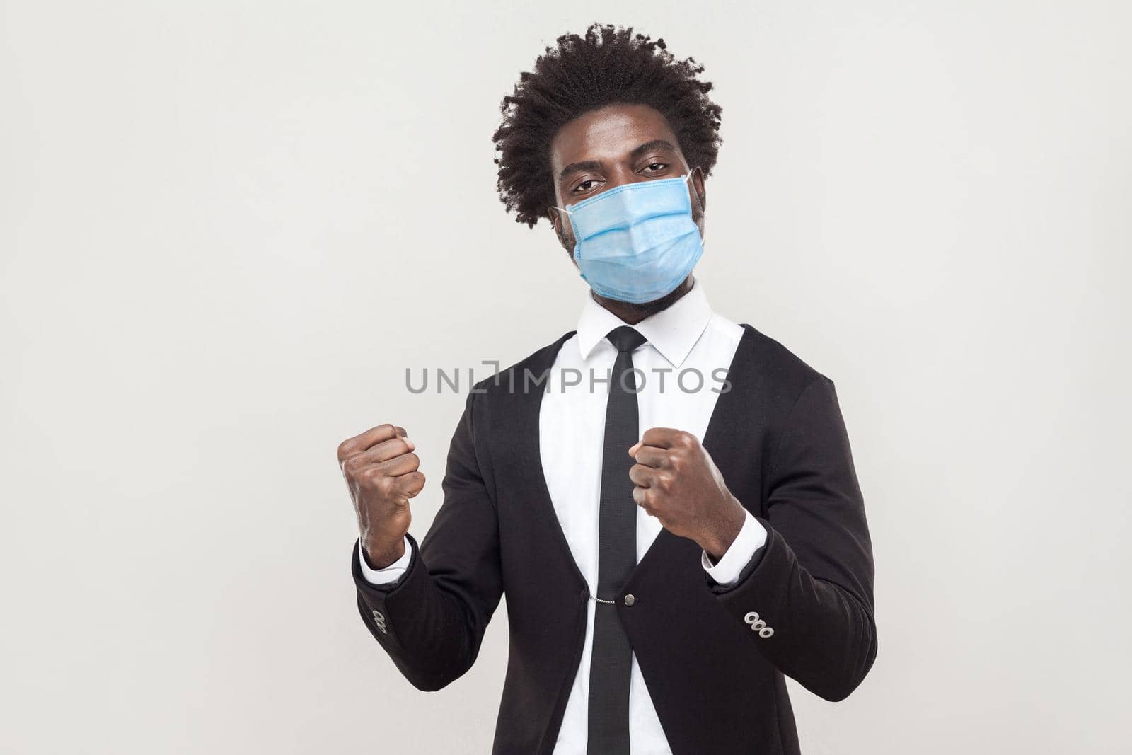 Yes, I win! Portrait of proud young handsome worker man wearing black suit with surgical medical mask standing with fists and celebrating his victory. indoor studio shot isolated on gray background.