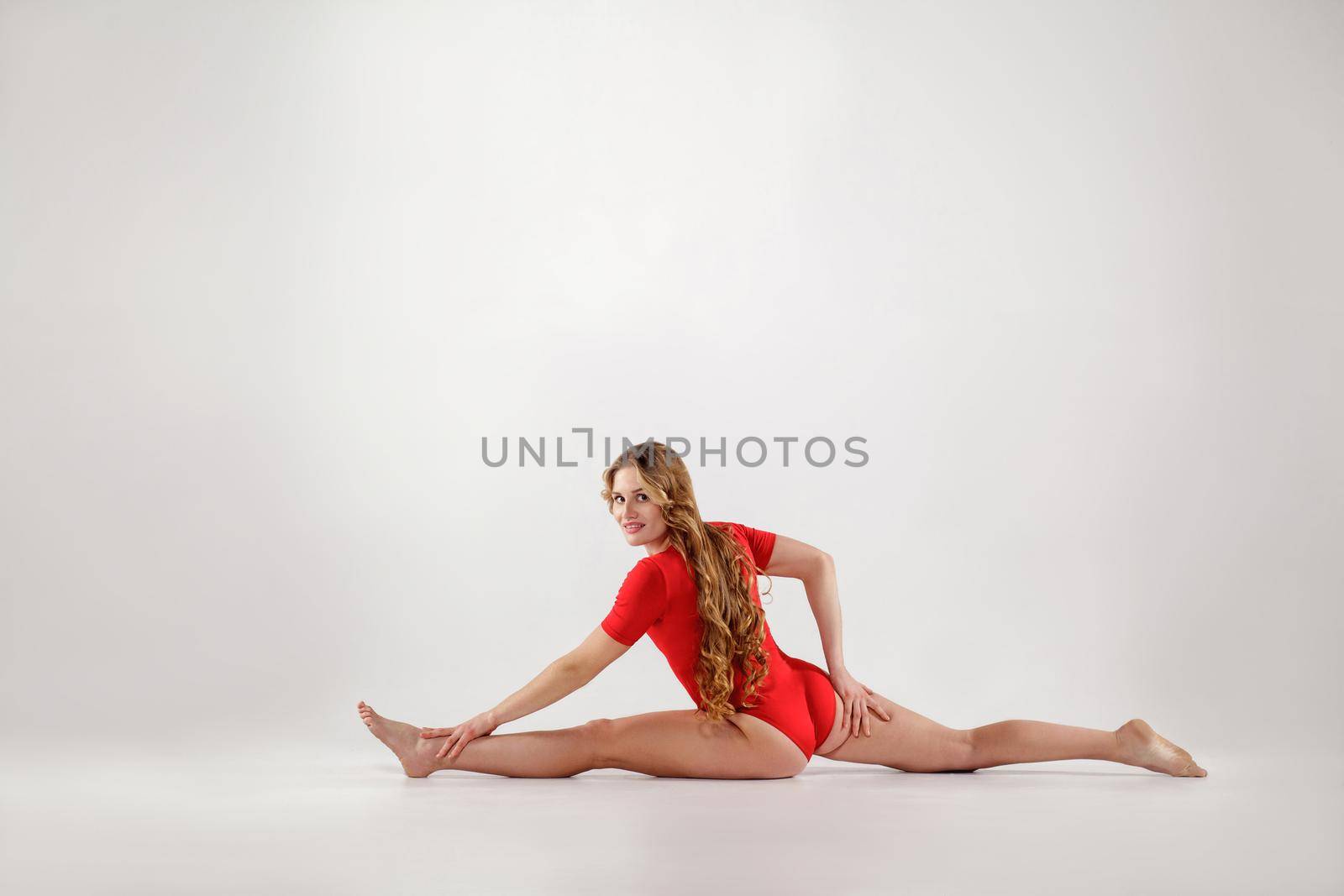 young athletic blonde woman in red leotard sitting on string. hanumanasana yoga pose.. isolated indoor studio shot on light gray background. healthy lifestyle and leisure activity concept.