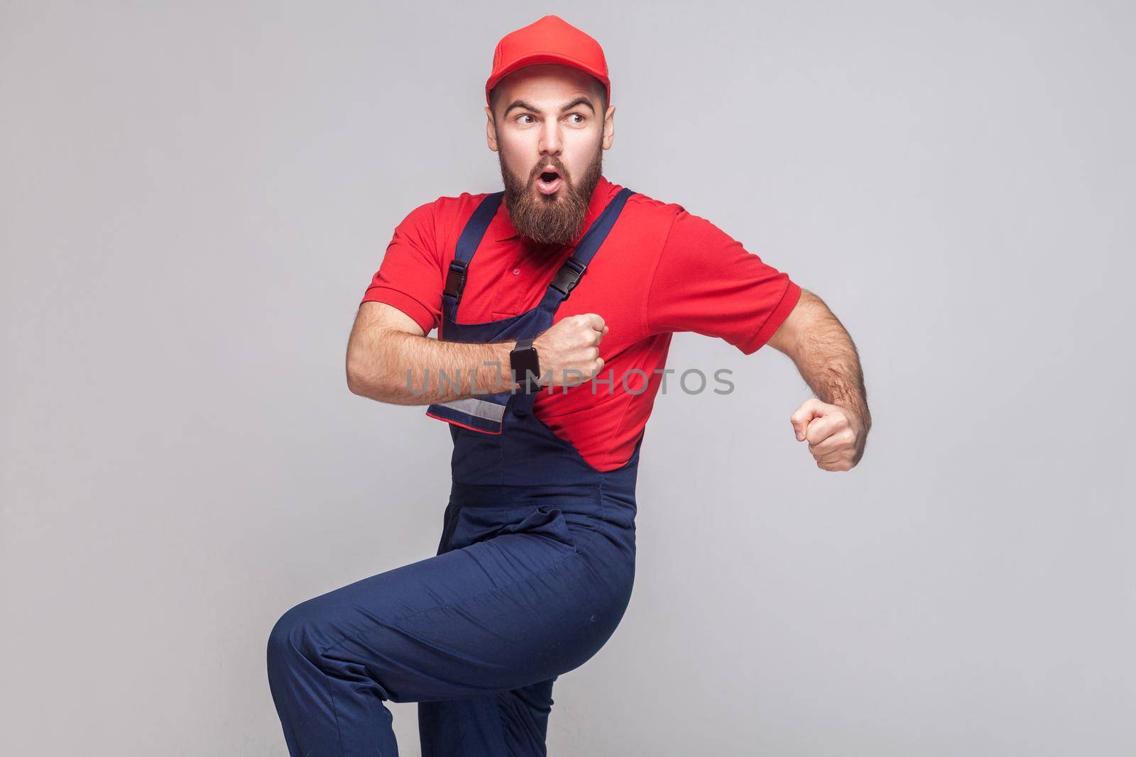 Hurry up! Young amazed handyman with beard in blue overall, red t-shirt and cap are late and starting to run for help on time. Grey background, indoor, studio shot, isolated, copy space