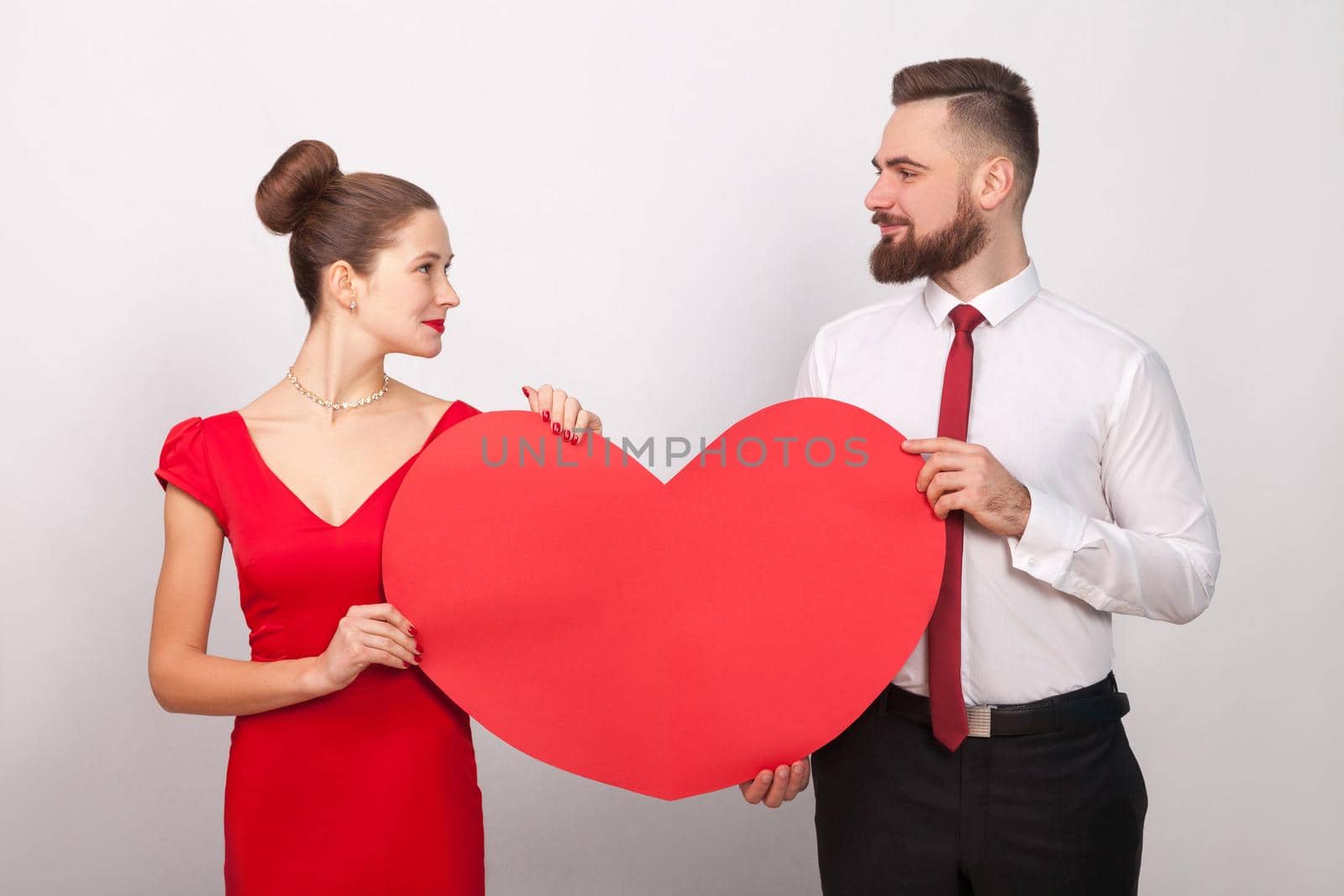 Beautiful couple looking each other, holding red heart, their love. Indoor, studio shot, isolated on gray background