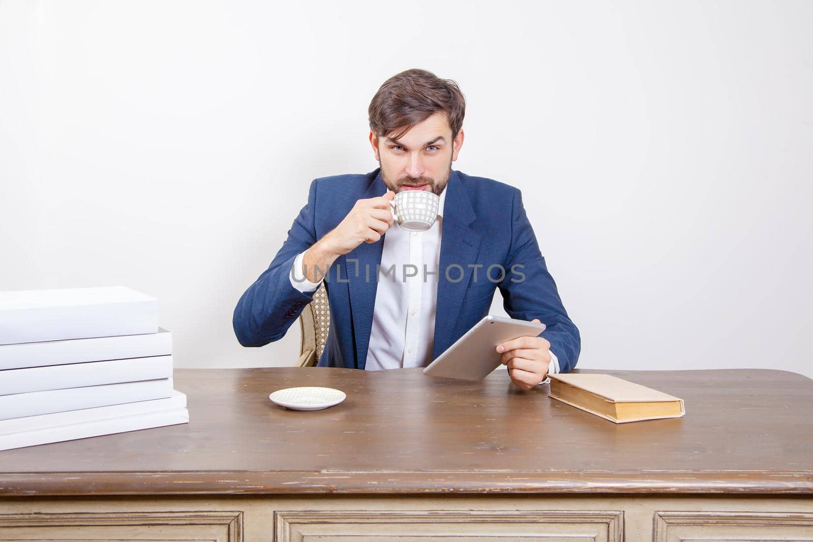 Technology, people and business concept - handsome man with beard and brown hair and blue suit and tablet pc computer and some books and cup drinking coffee or tea sitting in the office and looking at camera. .Isolated on white background. .