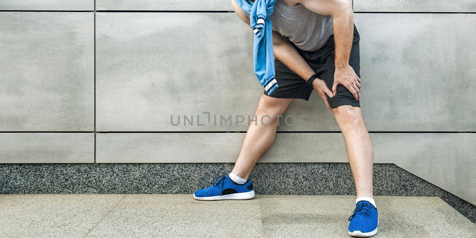 Knee ache and sport injury. Young man holding his painful knee and feeling bad. sport and healthcare lifestyle concept. cropped and standing on gray wall. outdoor shot, closeup.