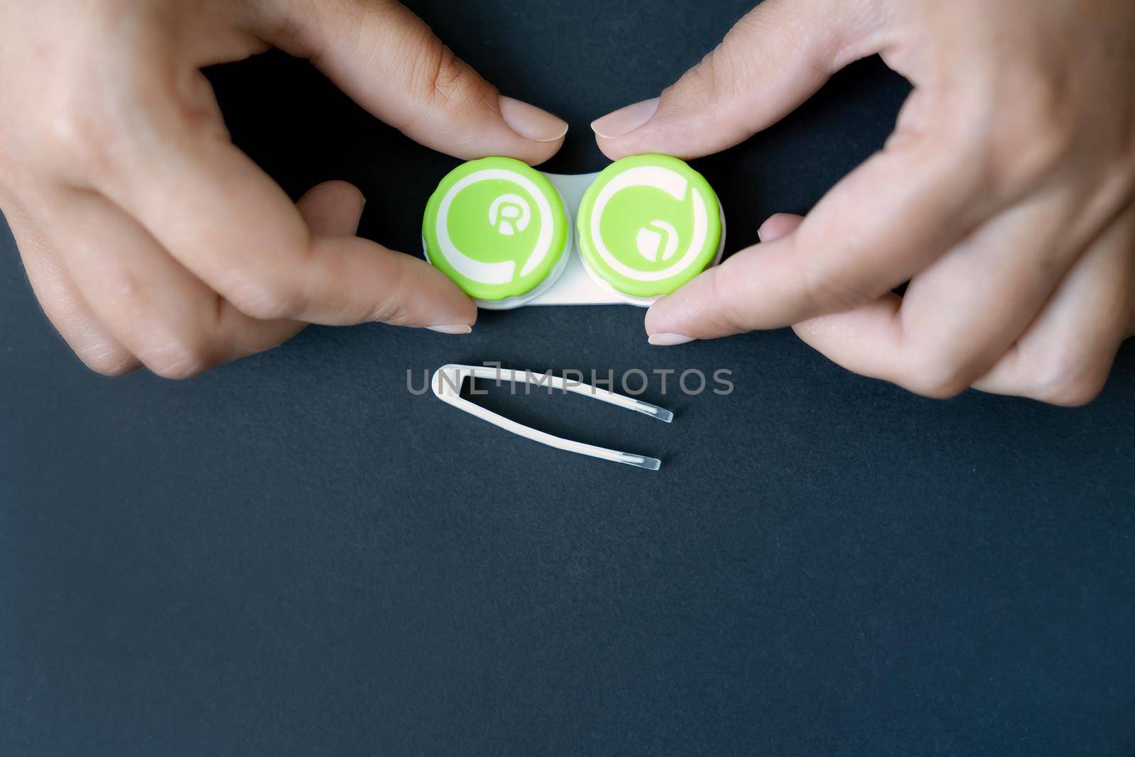 Girl's hands are holding container with lenses. Pair of tweezers for lenses lies next to it on black background. Close-up, top view, copy space