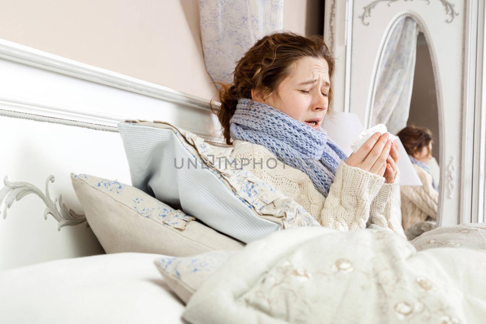 Sneezing beauty. Young sick woman with blue scarf on her neck sitting in bed and sneezing on her bed.