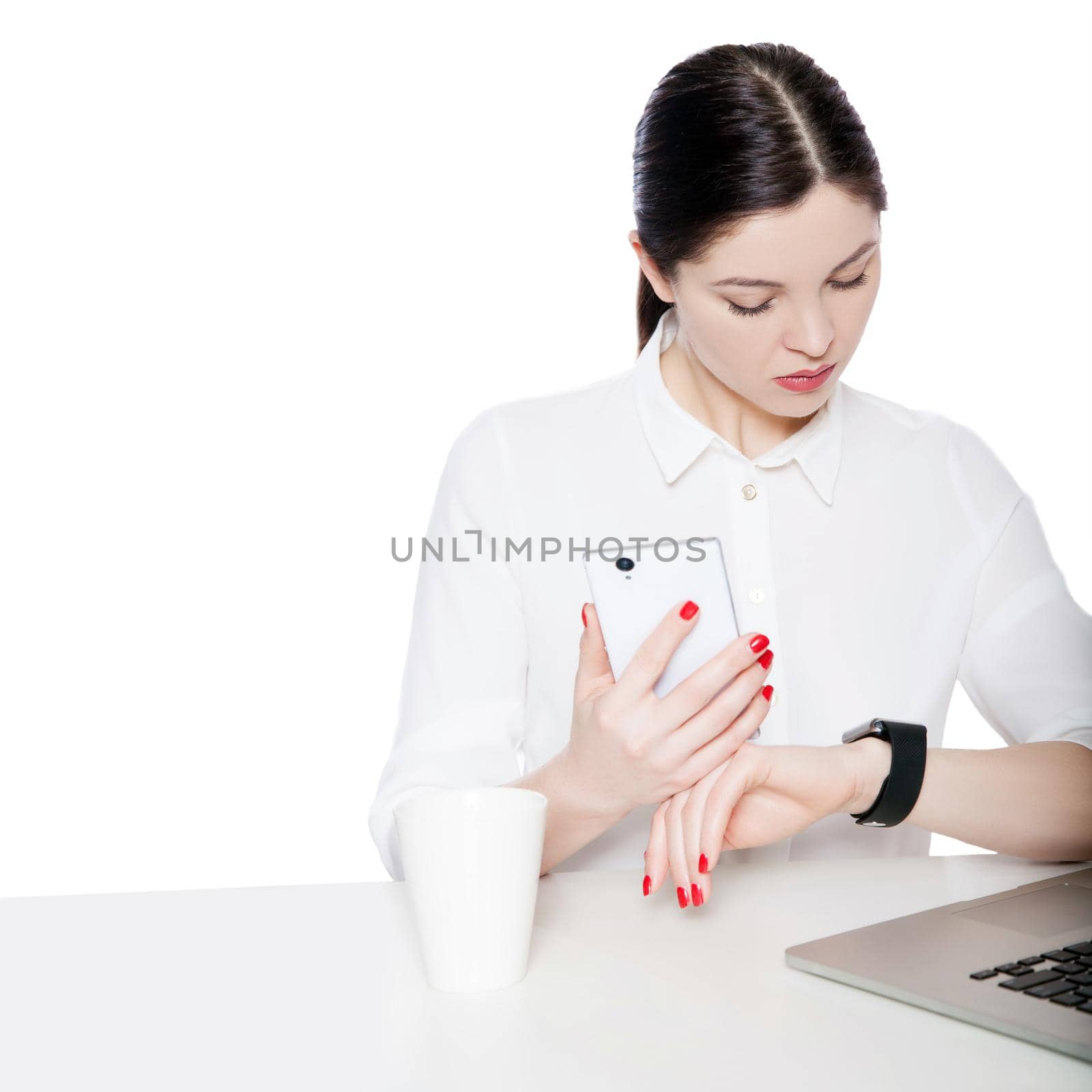 Portrait of attractive brunette businesswoman in white shirt sitting, holding smartphone mobile and looking at display of her smartwatch and reading something. studio shot isolated in white background