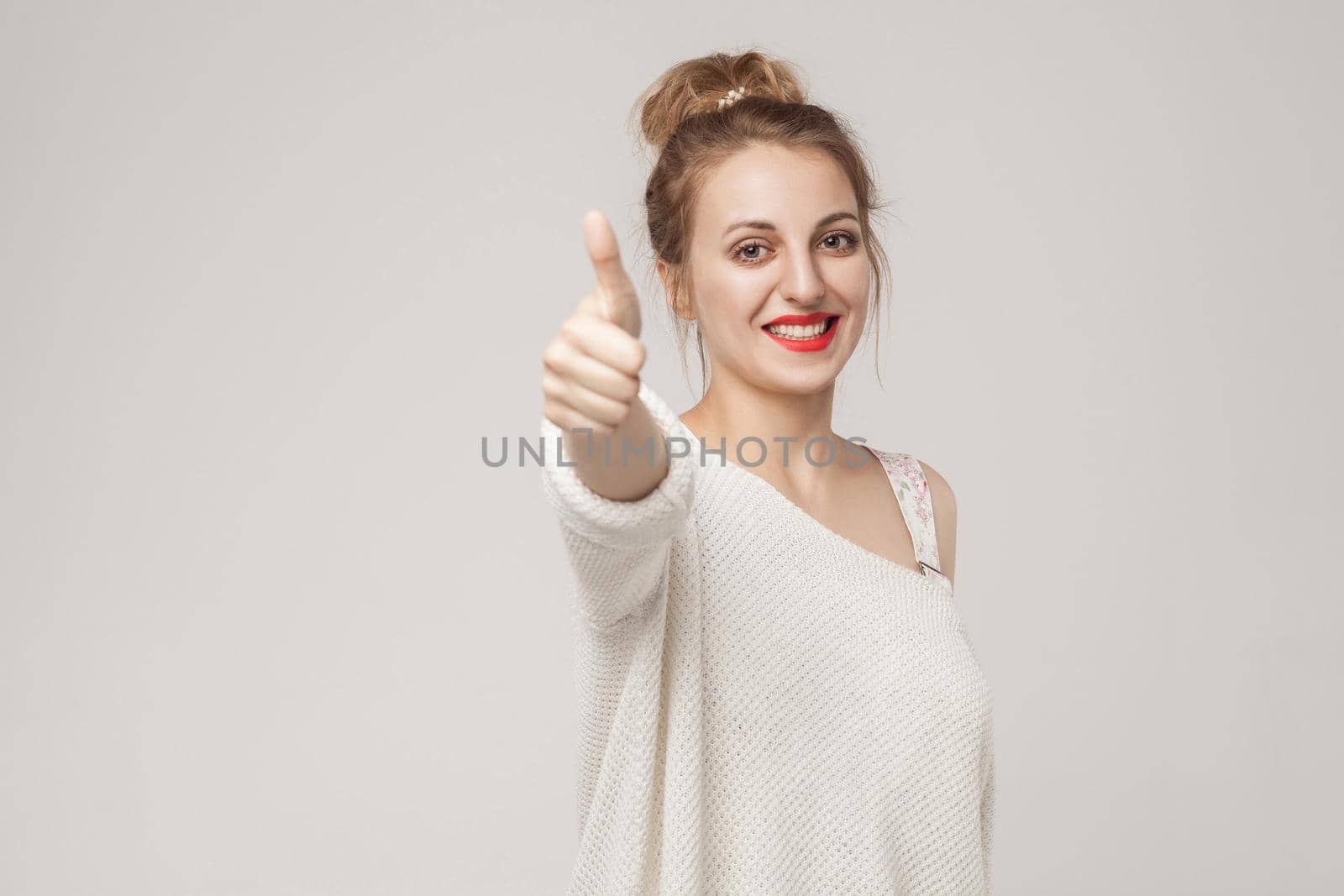 Young adult woman thumbs up, looking at camera with toothy smile. Studio shot, isolated on gray background