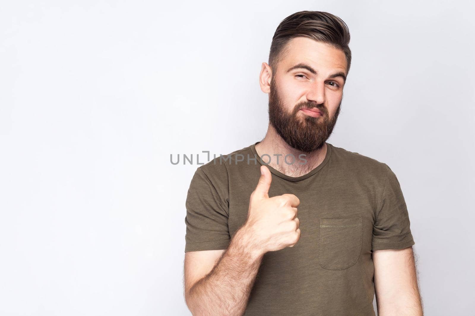 Portrait of satisfied bearded man with thumbs up and dark green t shirt against light gray background. studio shot. .