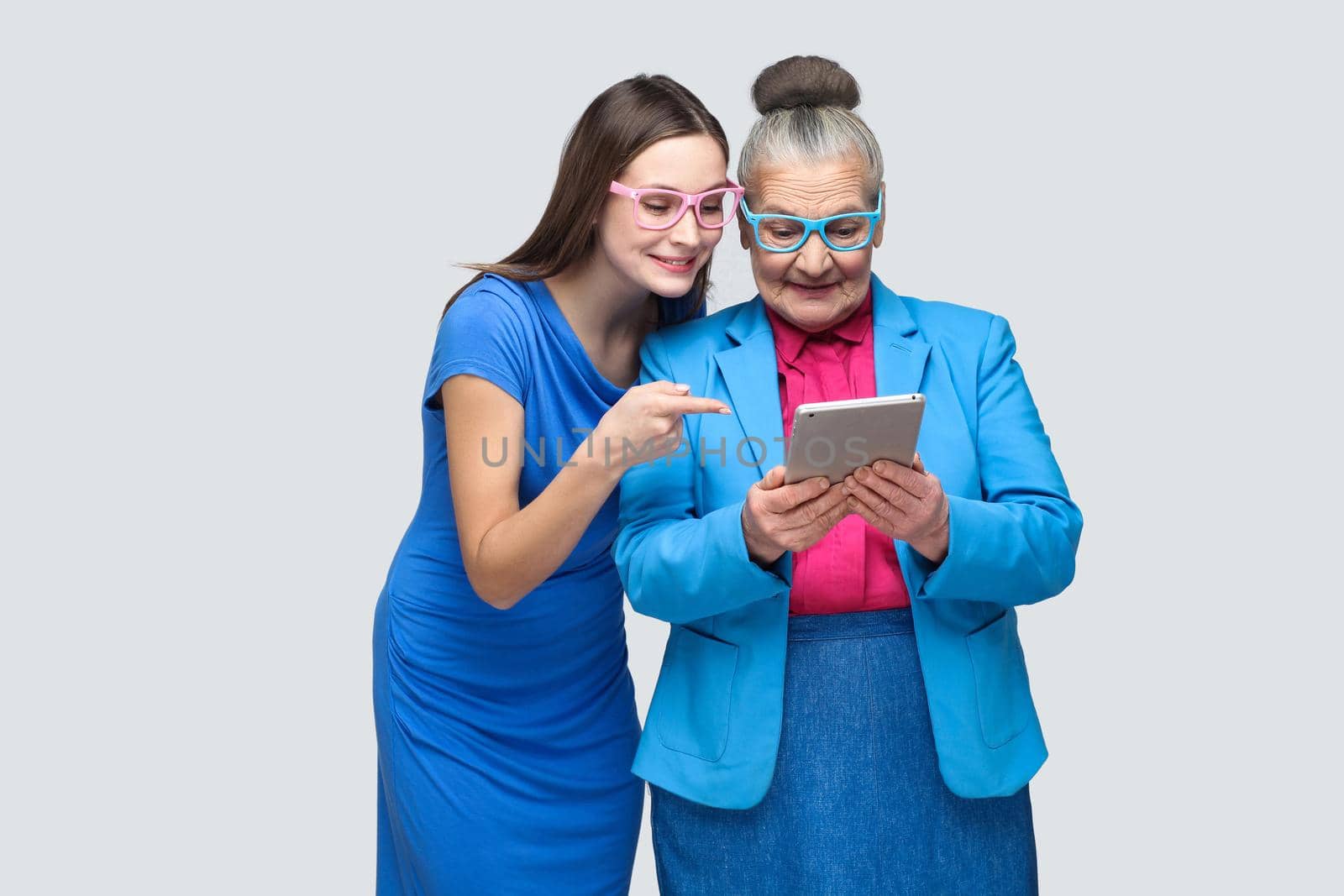 Young daughter in blue dress showing, pointing finger at tablet display in hands of aged woman. family relations between granddaughter and grandmother. indoor, studio shot, isolated on gray background