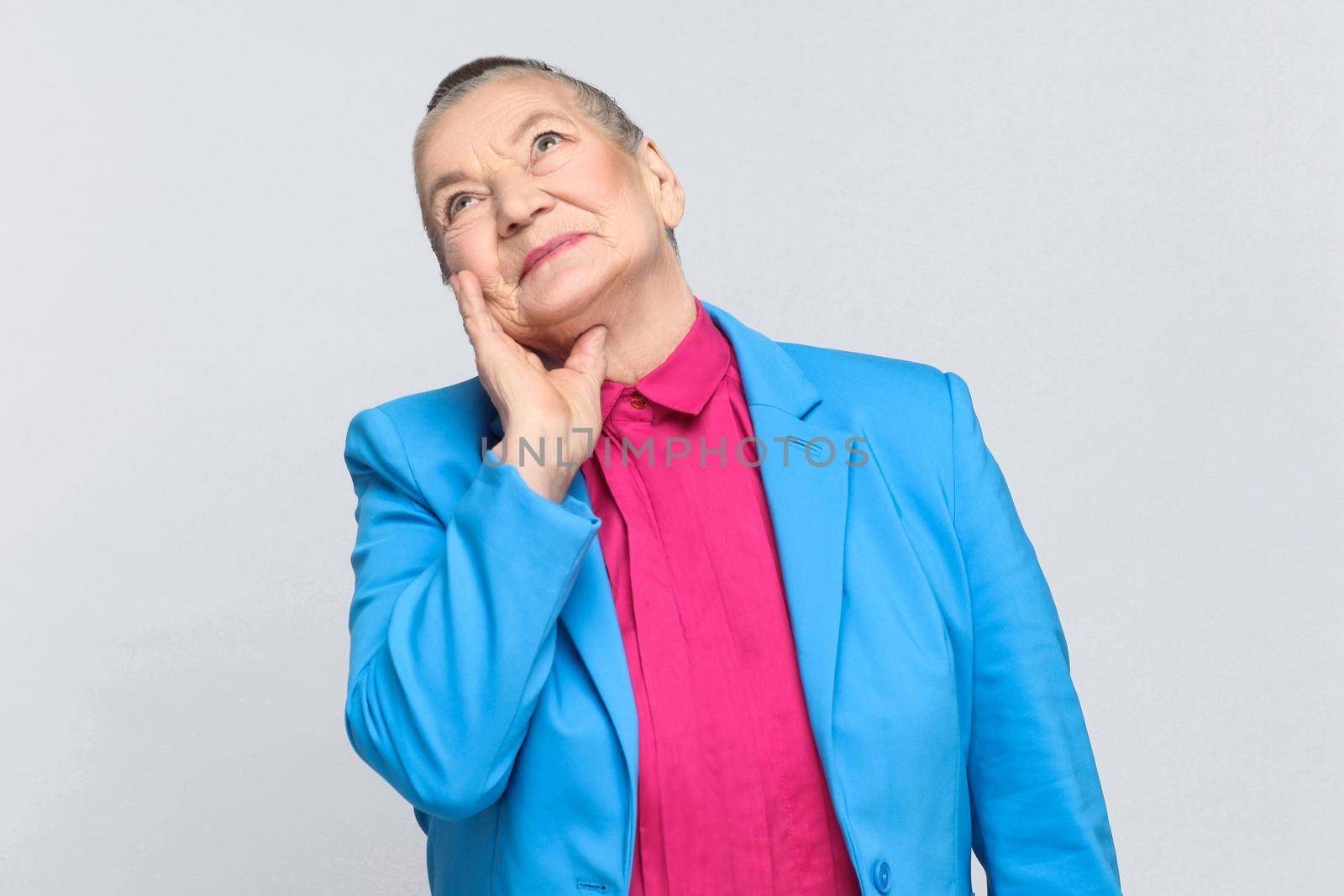 dreaming and imagination of future. Emotion and feelings handsome expressive grandmother with blue suit and pink shirt standing with collected bun gray hair. Studio shot, isolated on gray background