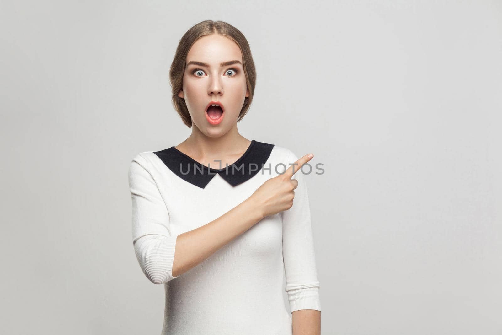 Boss pointing finger at copy space, and looking at camera with shocked face and big eyes. Studio shot, gray background