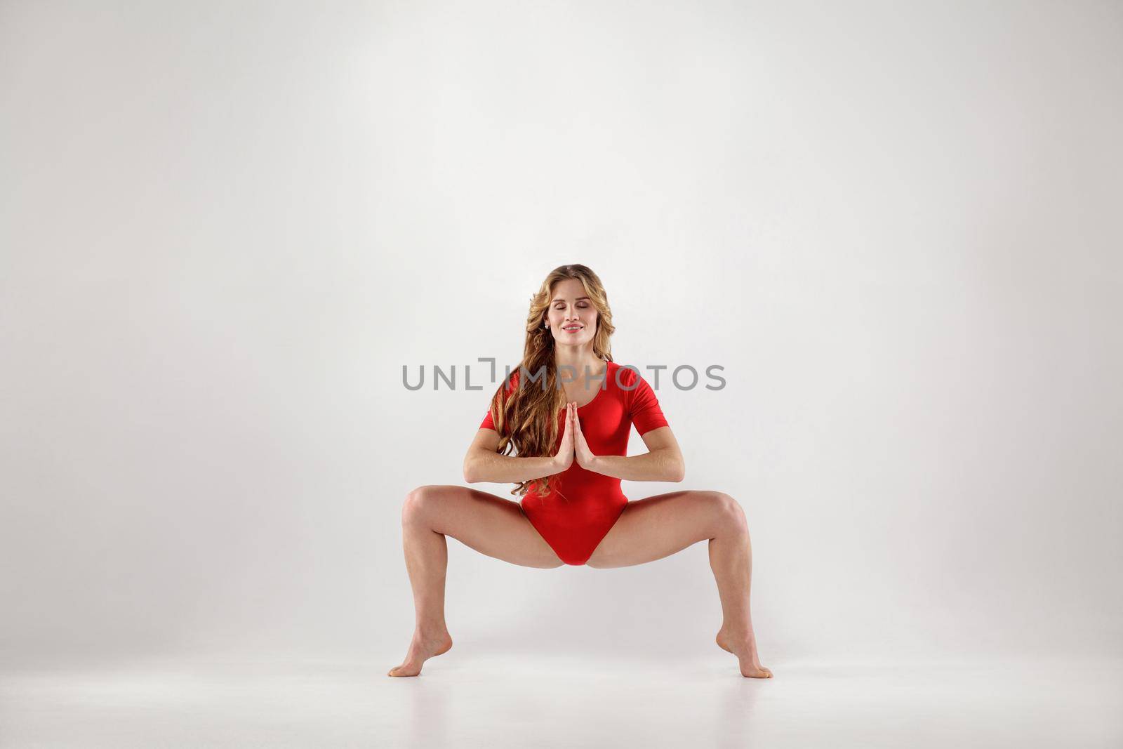 attractive athletic curly blonde woman with makeup in red leotard doing yoga pose .isolated indoor studio shot on light gray background. healthy lifestyle and leisure activity concept.