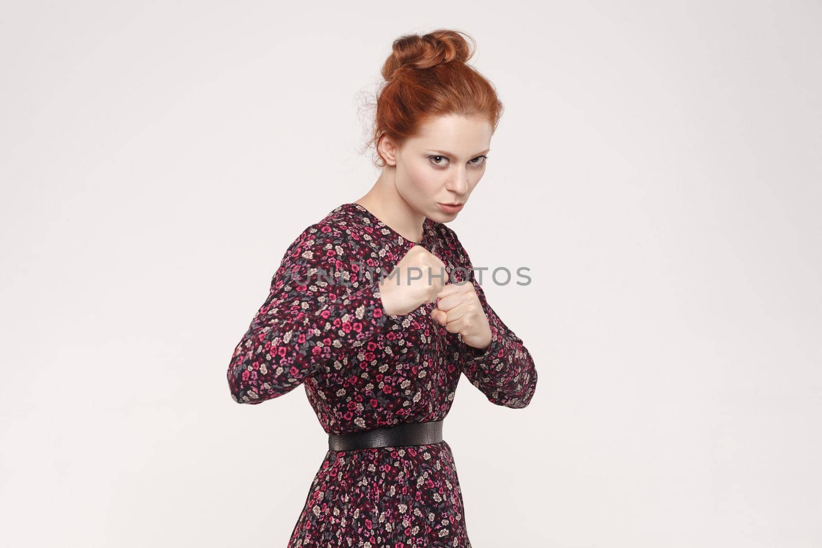 Ready to fight! Aggressive redhead women with bun hair looking at camera with seriously face. Indoor, isolated on gray background