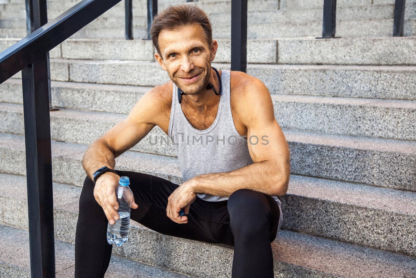 Happy smily sportsman resting and holding bottle of water, sitting on steps looking at camera and smiling . fitness, sport, exercising and people healthy lifestyle concept.
