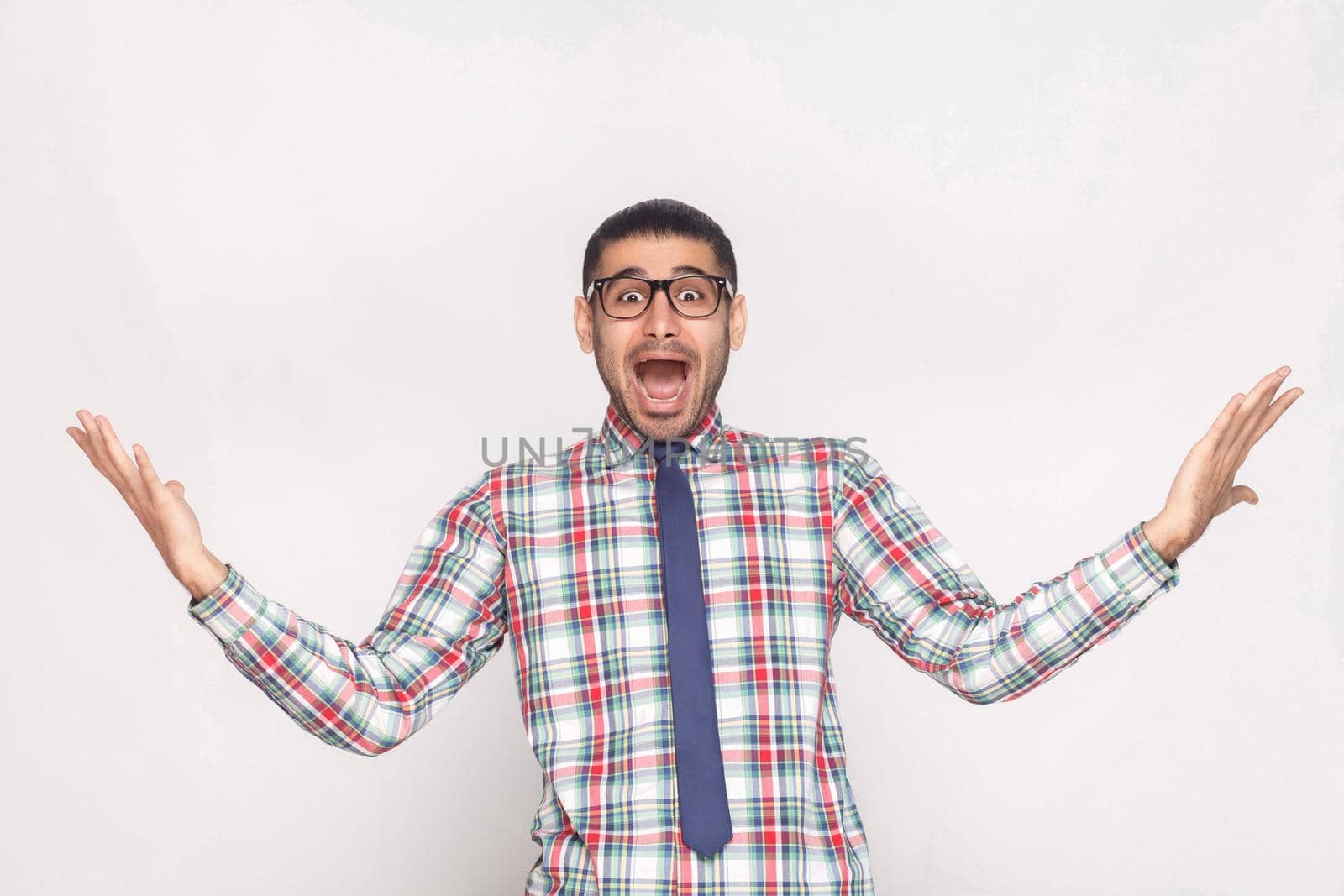 Amazed or shocked bearded businessman in checkered shirt, blue tie and eyeglasses standing, raised hand and looking at camera with surprised open mouth. studio shot, isolated on light grey background.