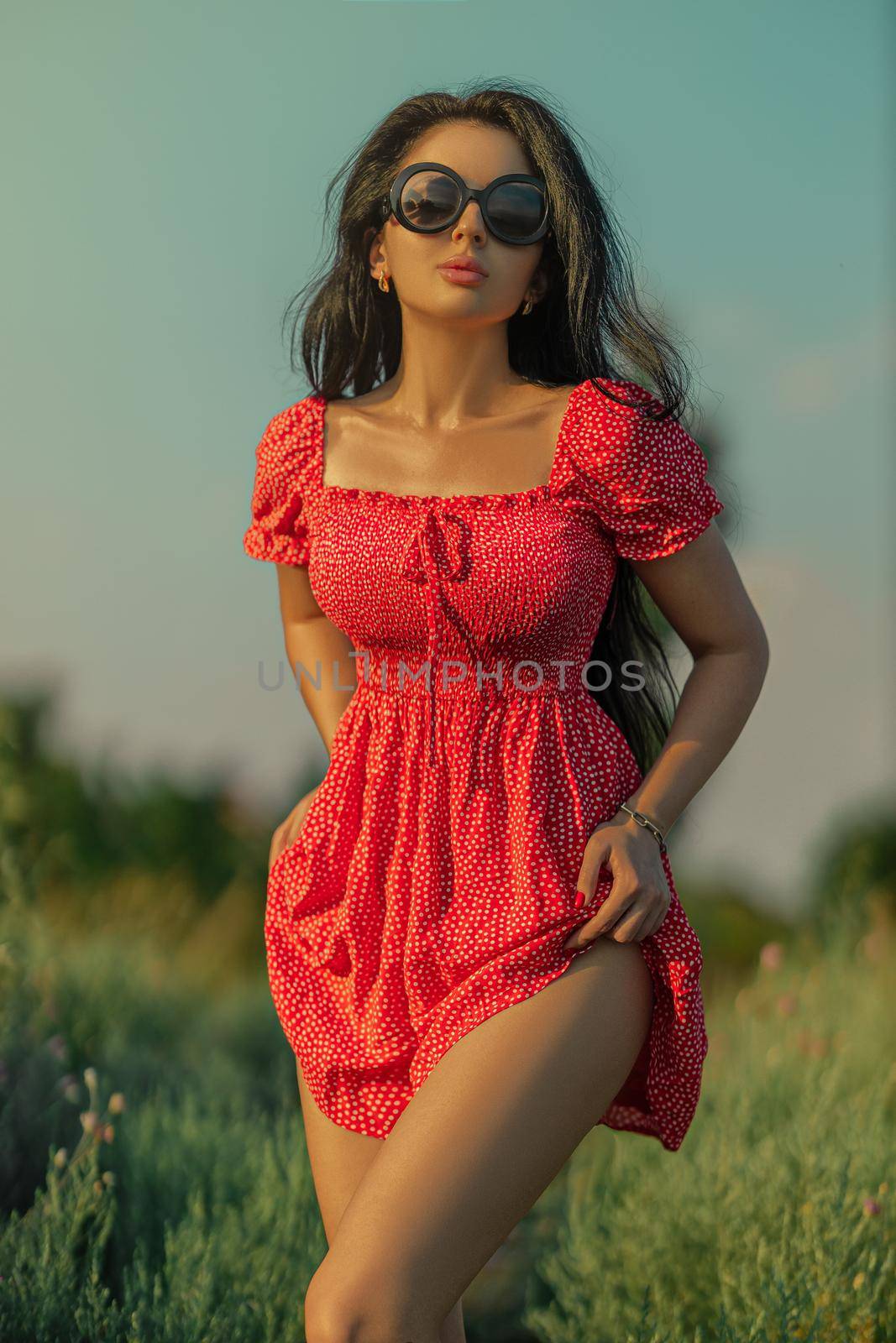Sexy brunette girl in a red dress with polka dots posing in a meadow among wild herbs, flowers and trees on a summer sunny day
