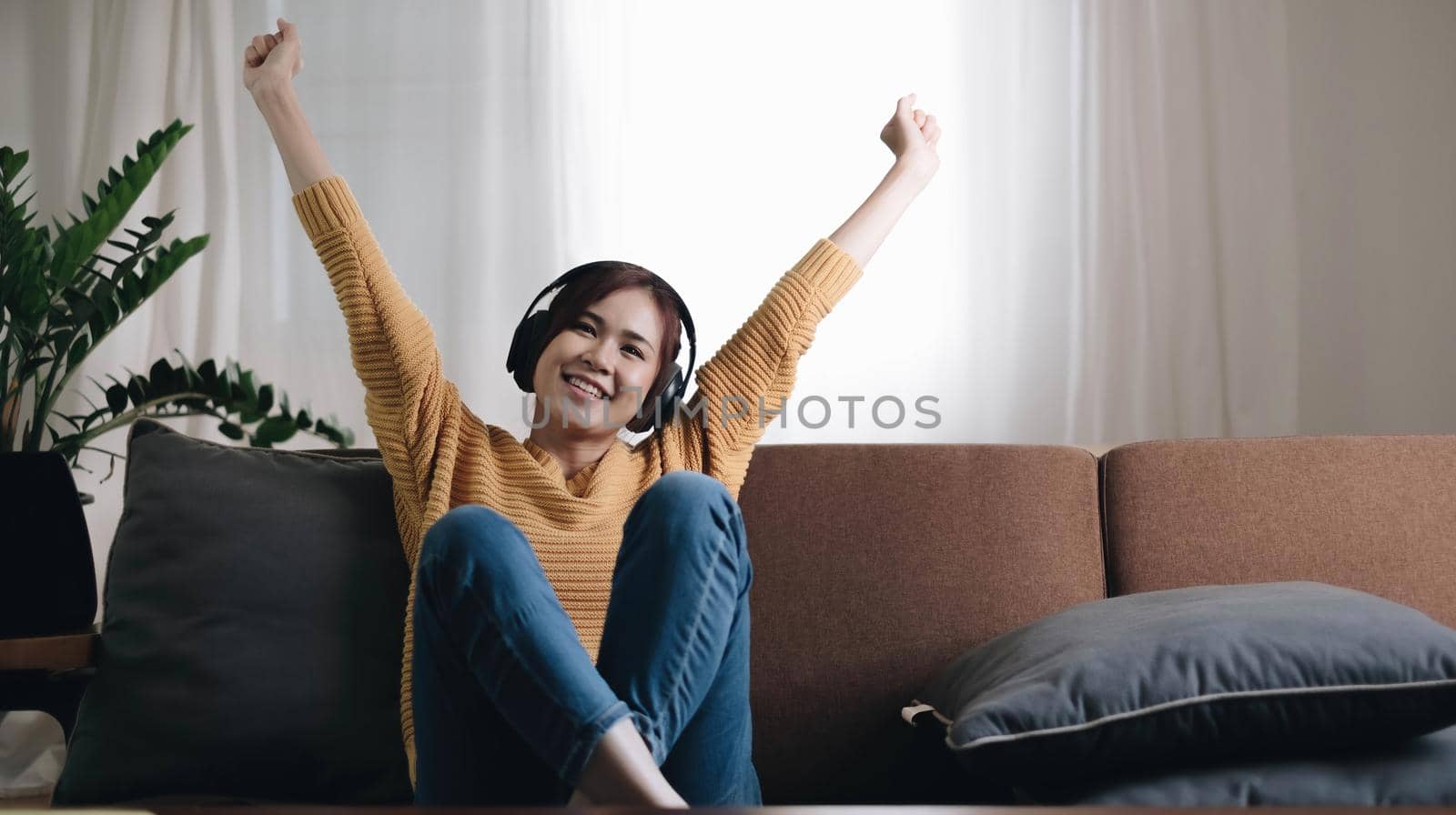 Being energetic. Pretty overjoyed young woman laughing and listening to music and wearing headphones while sit on the sofa and wearing a yellow sweater by wichayada