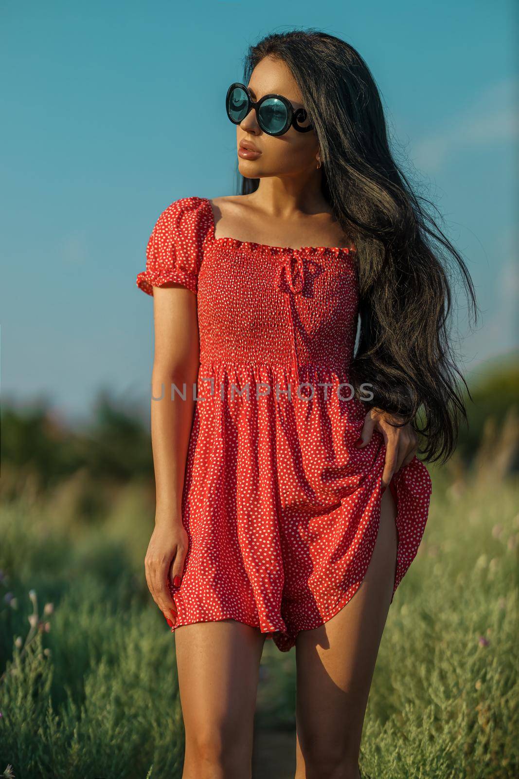 Tanned sexy brunette girl in a red dress with small polka dots posing barefoot in a meadow among wild herbs, flowers and trees on a summer sunny day
