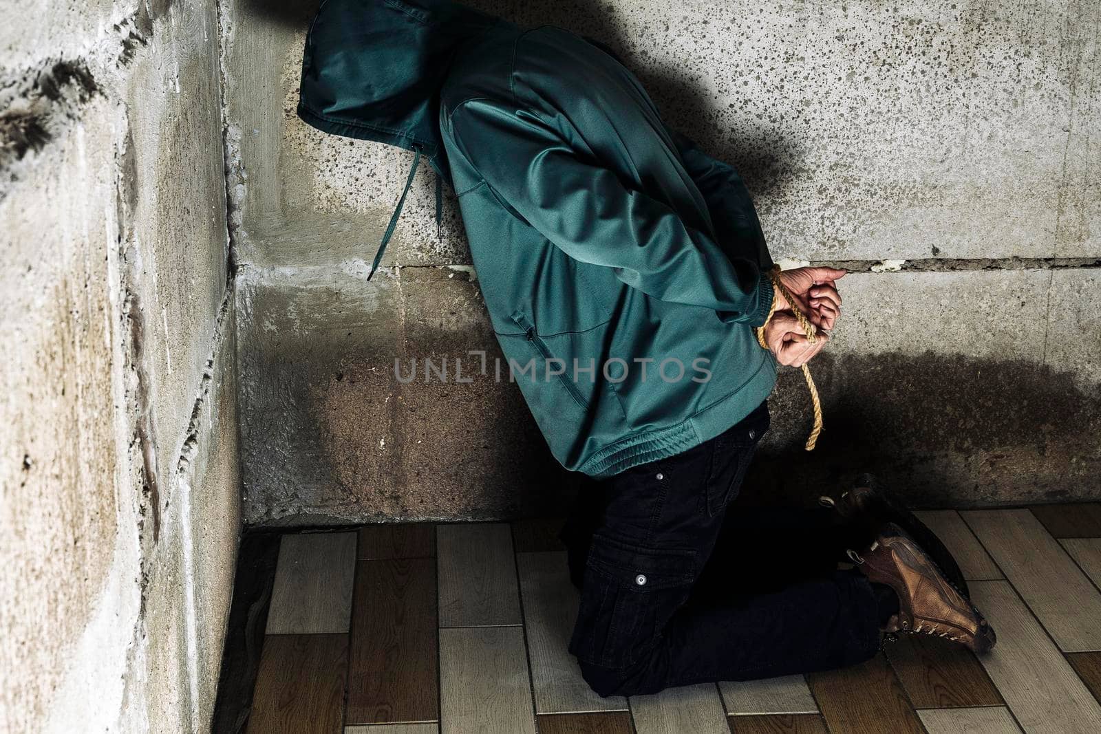 an unrecognizable man in a hood is captured in the basement bound by a rope hand and foot . The kidnapped man is tied up, sitting in the basement. Hands and feet are tied with rope. kneeling on the floor