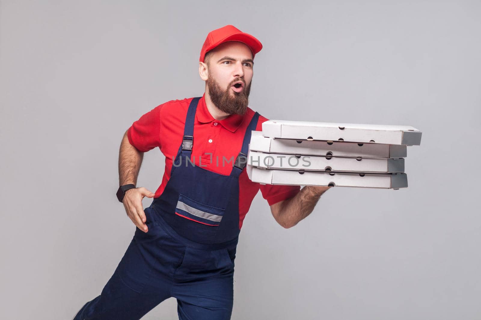 Hurry up! Young amazed delivery man with beard in blue uniform and red t-shirt holding stack pizza boxes and running to delivery on grey background. Indoor, studio shot, isolated, copy space.