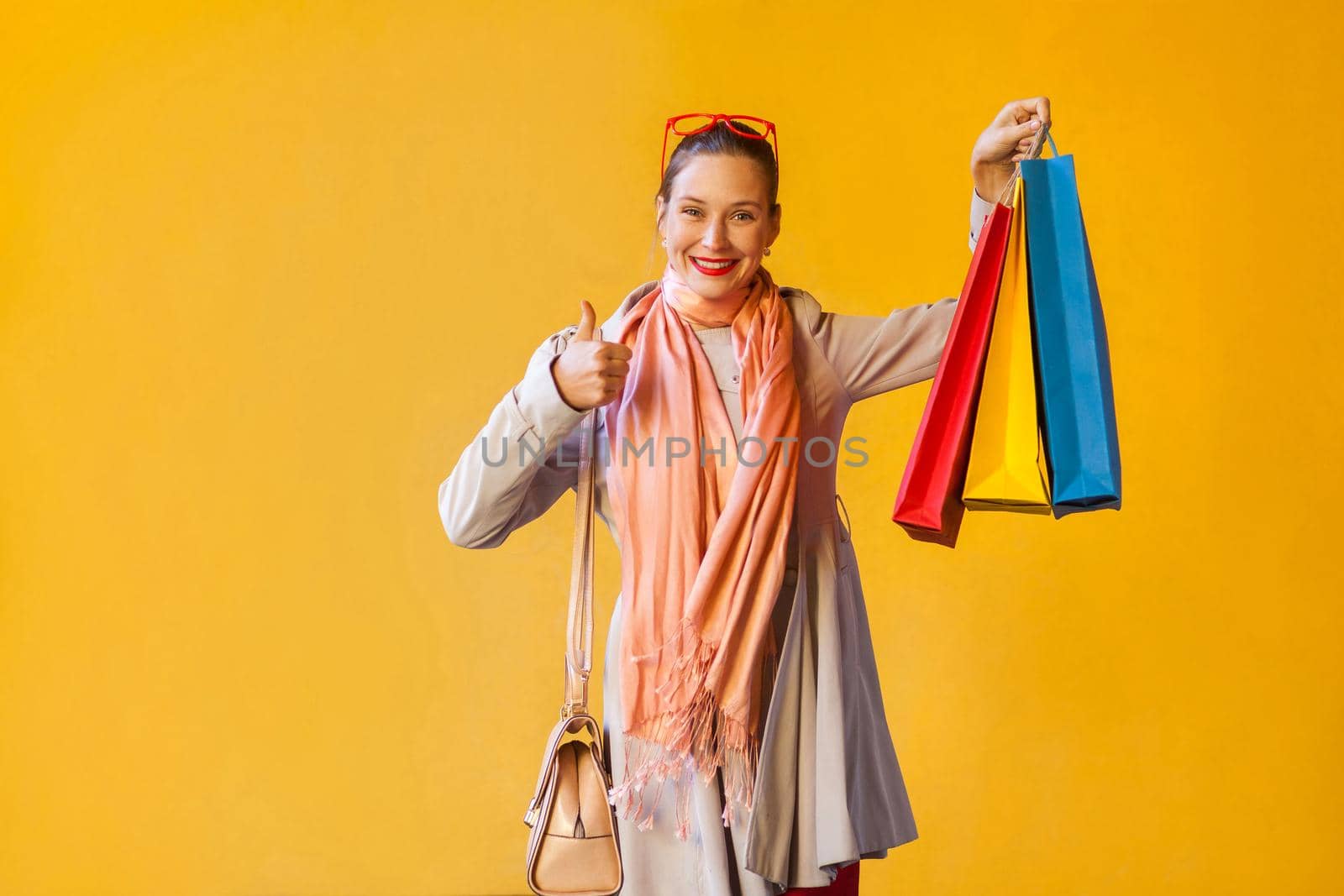 Happy cheerful young adult girl with freckles, bun hair, looking at camera, thumbs up finger, holding many shopping bag, having joyful look, enjoying good day and free time indoors. Studio shot