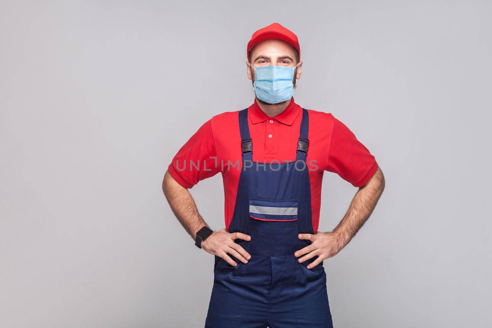 Portrait of young man with surgical medical mask in blue overall, red t-shirt and cap standing and holding hands on waist and looking at camera, indoor, studio shot, isolated on gray background