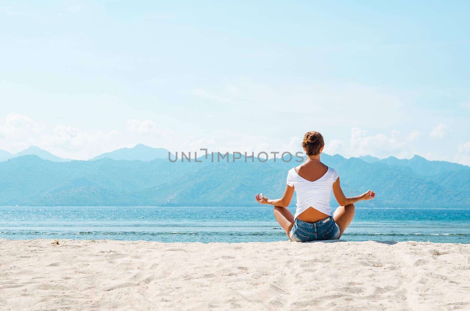 A young woman meditates in a pose of yoga on a sandy beach overlooking the blue ocean and the mountains. Stock image.