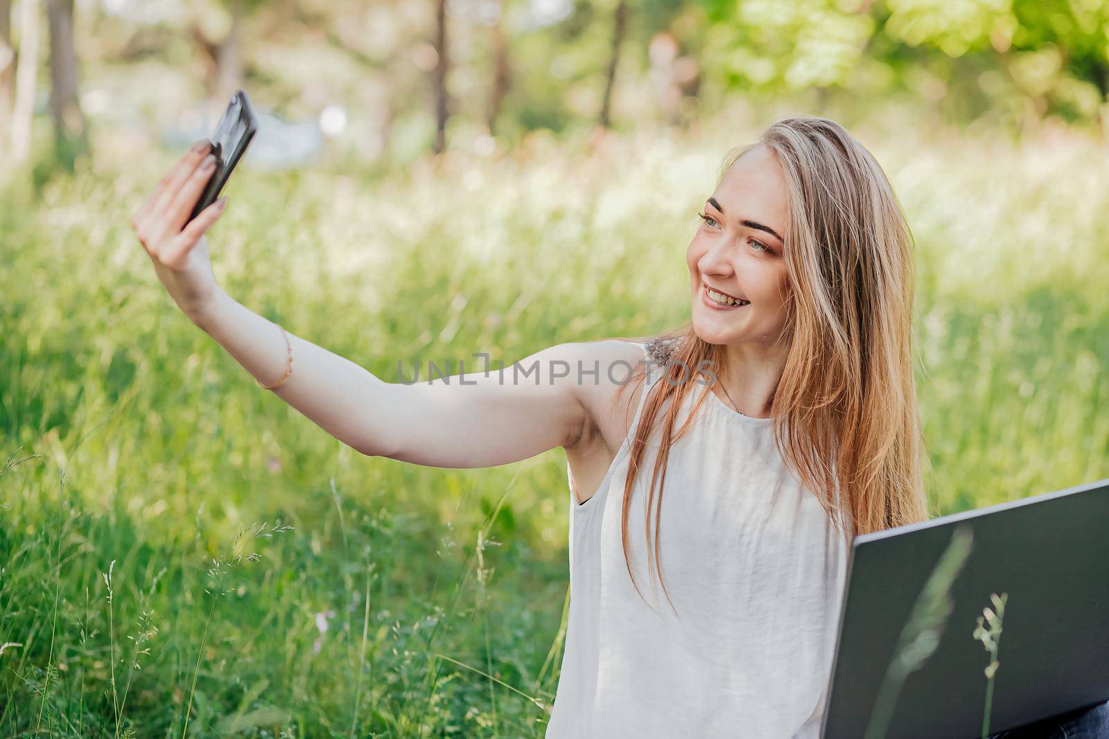 girl sits outdoors and works at a laptop. makes a selfie on the phone. freelance. selfeducation. the concept of remote learning and outdoor work. by Anyatachka