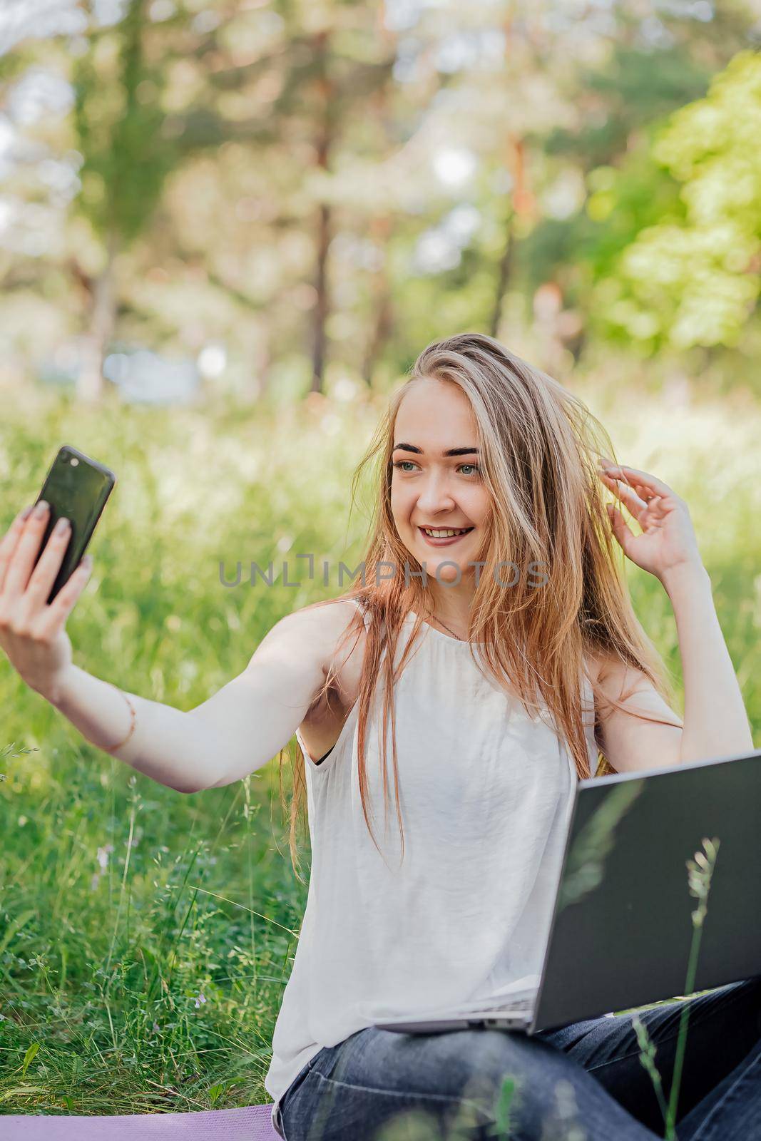 girl sits outdoors and works at a laptop. makes a selfie on the phone. freelance. selfeducation. the concept of remote learning and outdoor work